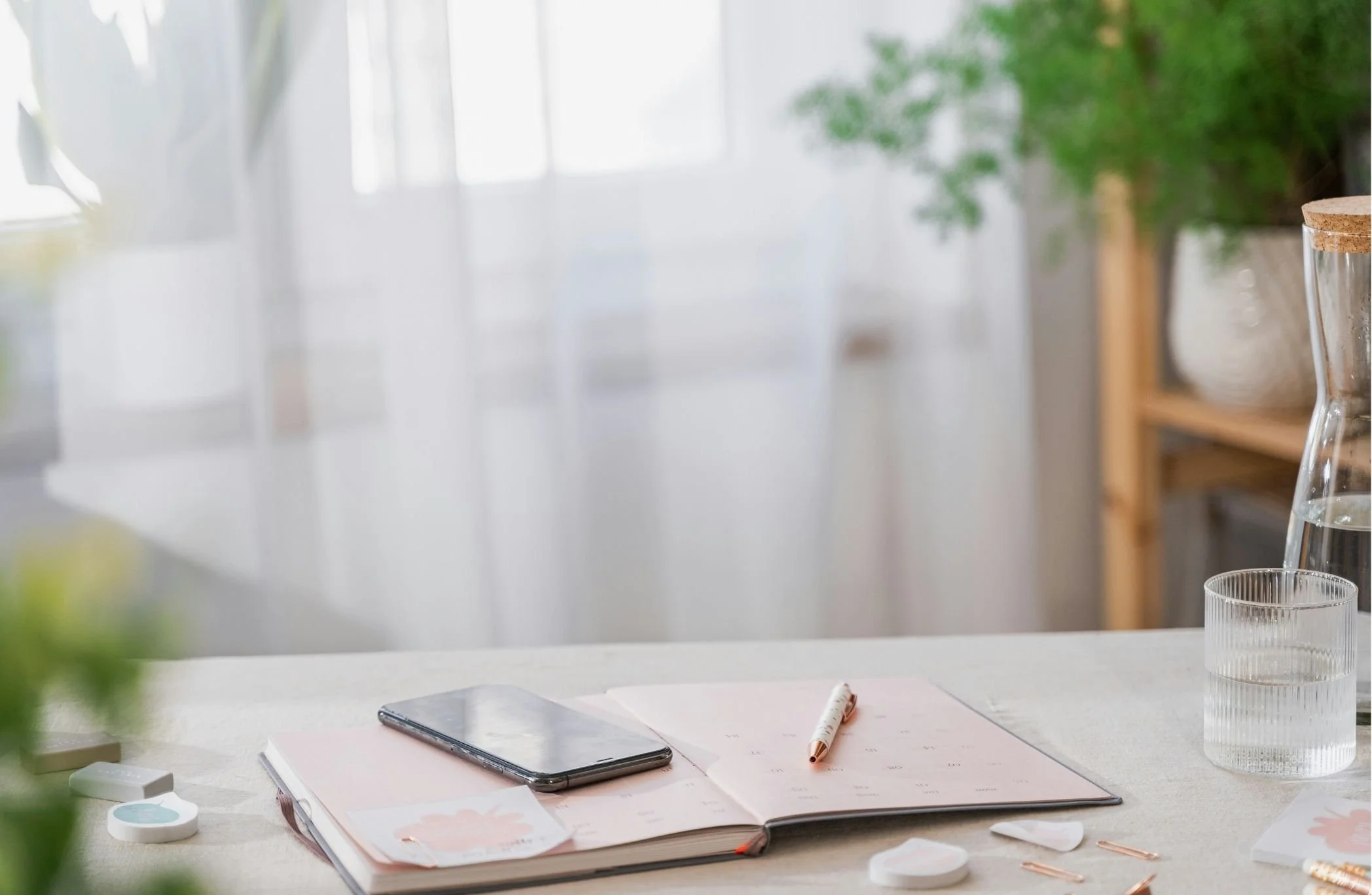 A workspace table with an open planner, a smartphone, and a pen. There are various office supplies around, with a blurred background of window and greenery.