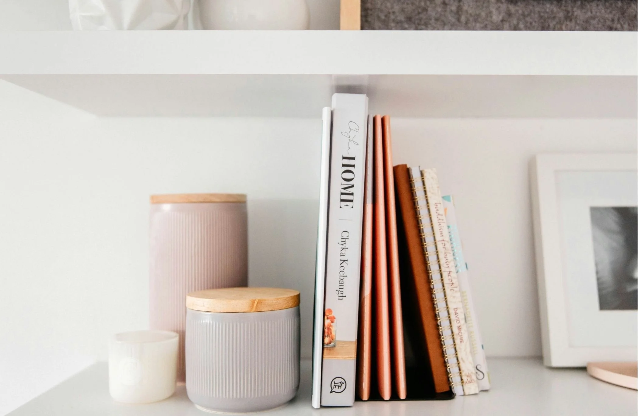 Shelf with white and orange-colored books, a ceramic container with wooden lid, a pink ribbed cup, and a white container, against a white wall.