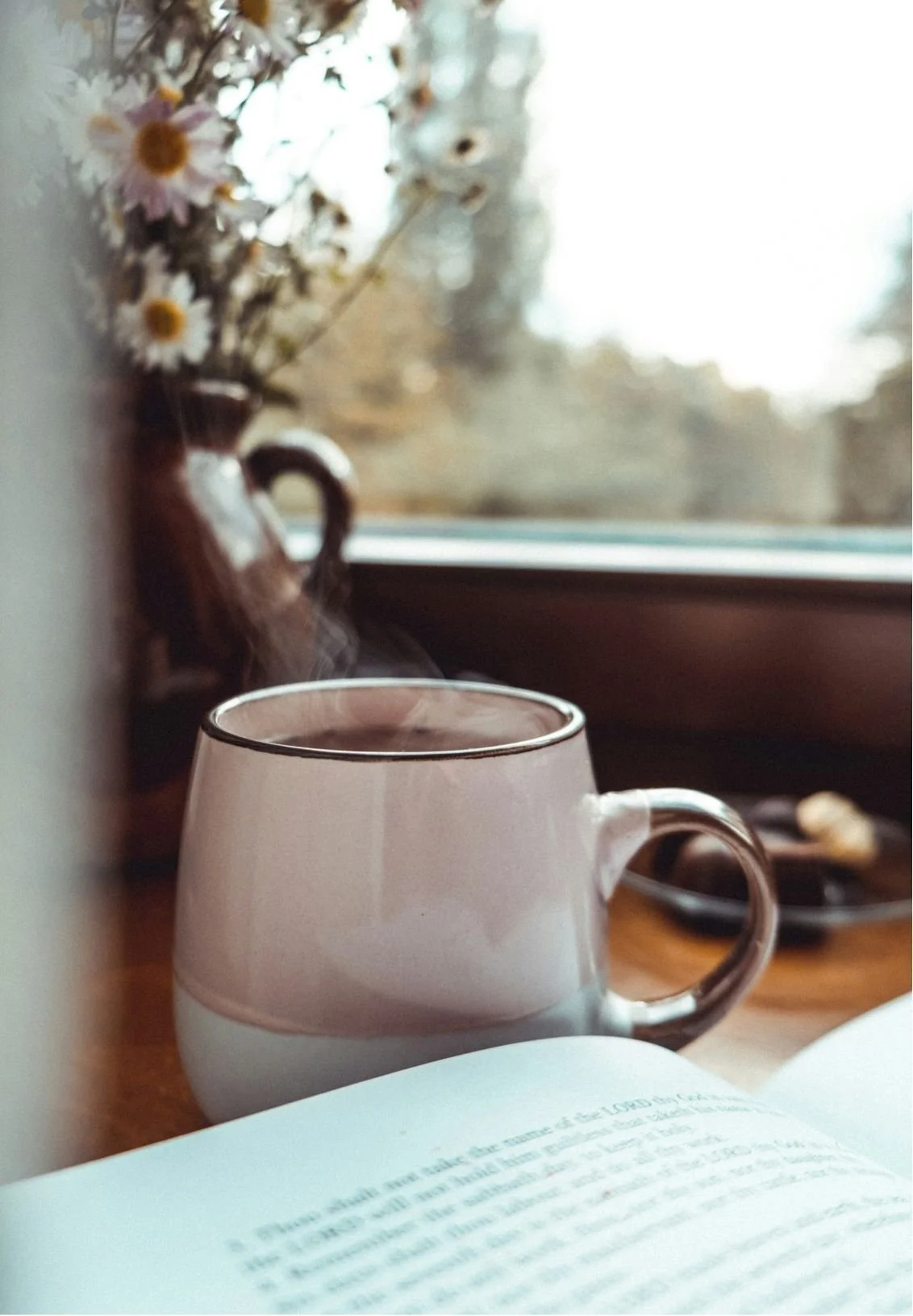 A steaming cup of coffee on a wooden surface next to an open book, with a vase of dried flowers and a window in the background.