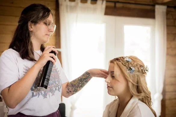 A woman with glasses and tattoos is applying makeup or setting spray to another woman with long blonde hair, wearing a floral hair accessory, in a room with wooden walls and white curtains.
