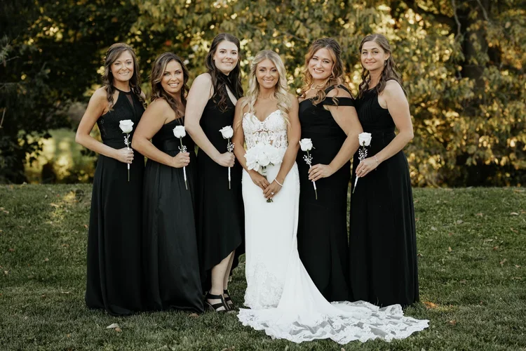 A bride in a white wedding dress holding a bouquet, surrounded by five women in black dresses, each holding a white rose, standing outdoors on grass with trees in the background.