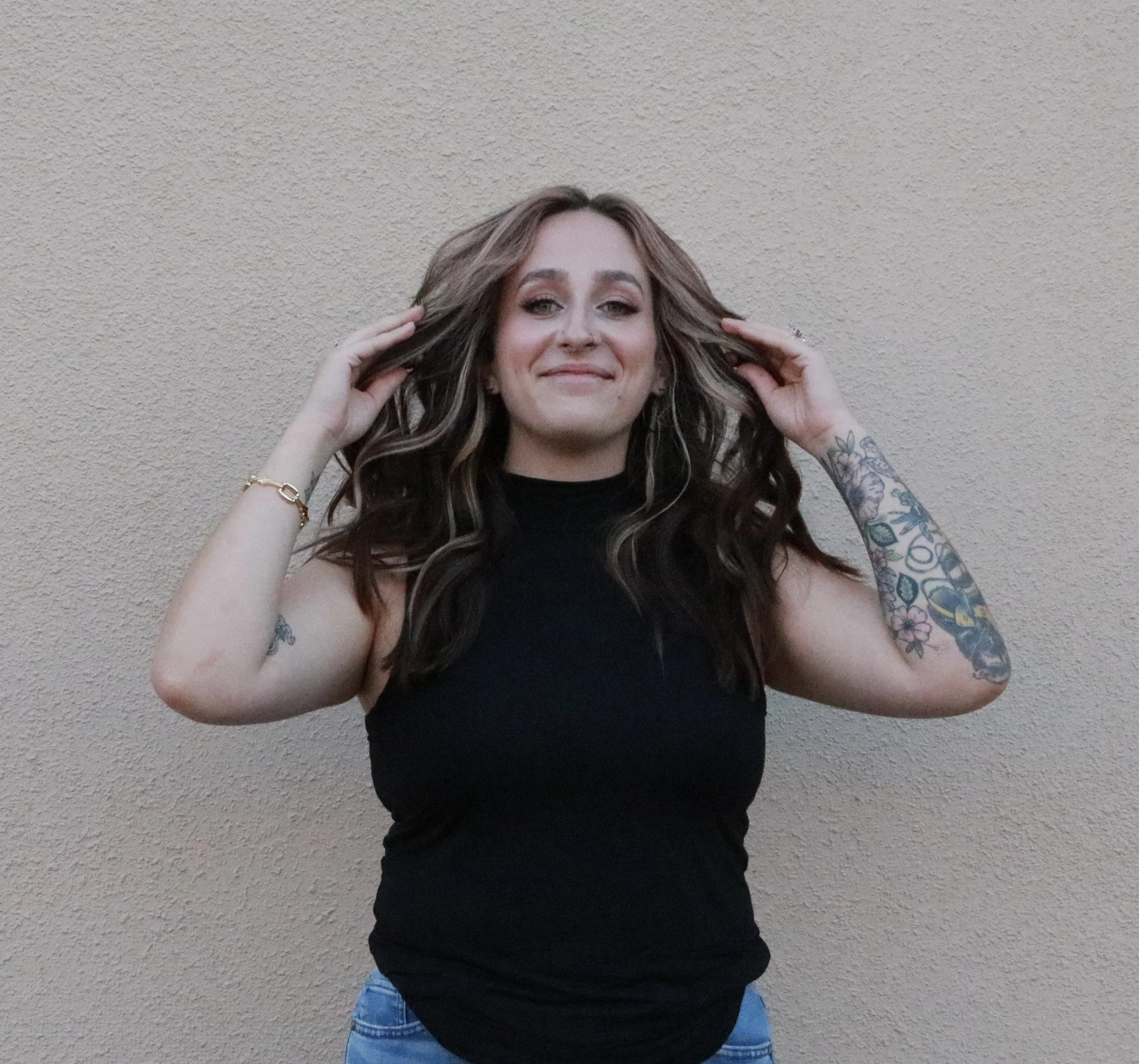 A woman with long, wavy brown hair and tattoos on her left arm, wearing a black shirt, standing against a beige textured wall. She is smiling and holding her hair with both hands, looking at the camera.