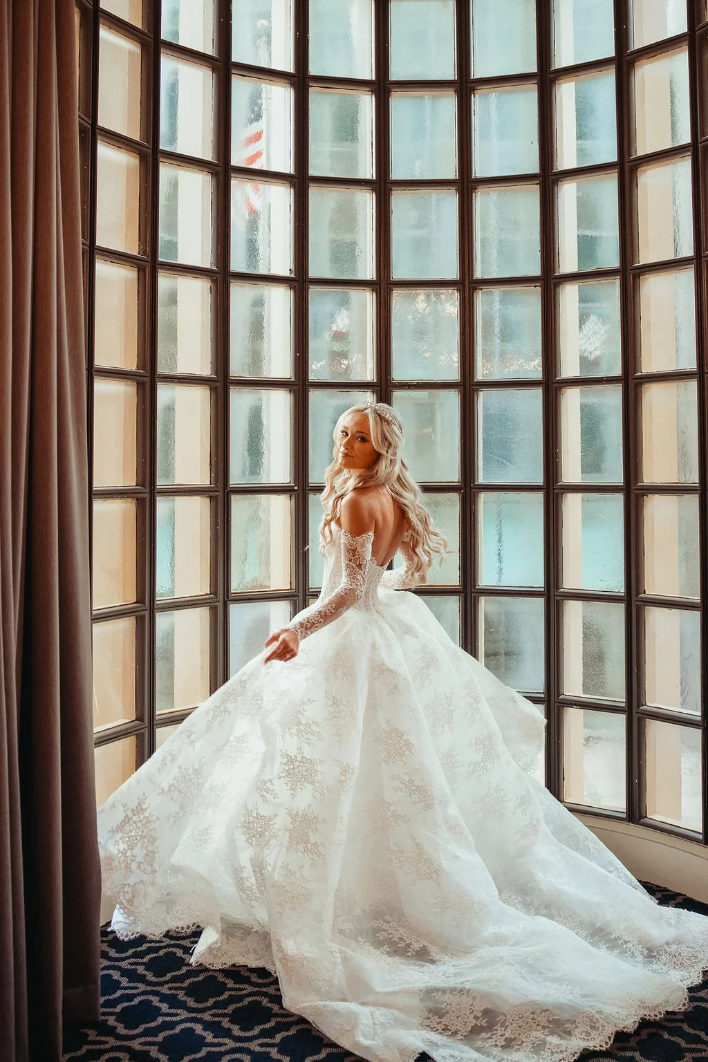 A bride in a white wedding gown with lace details stands near a large window with multiple panes. She has long, wavy blonde hair and is looking over her shoulder.