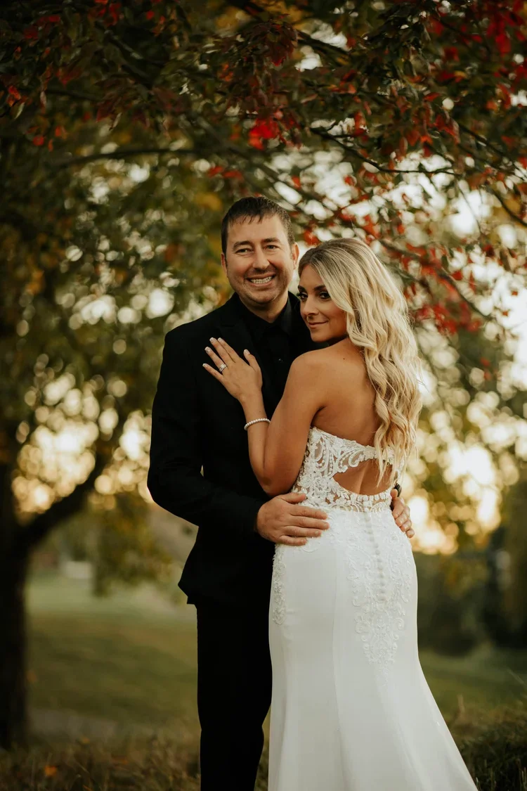 A newlywed couple standing outdoors under trees with fall foliage, the groom in a black suit and the bride in a white lace wedding gown, smiling and posing for a photo.