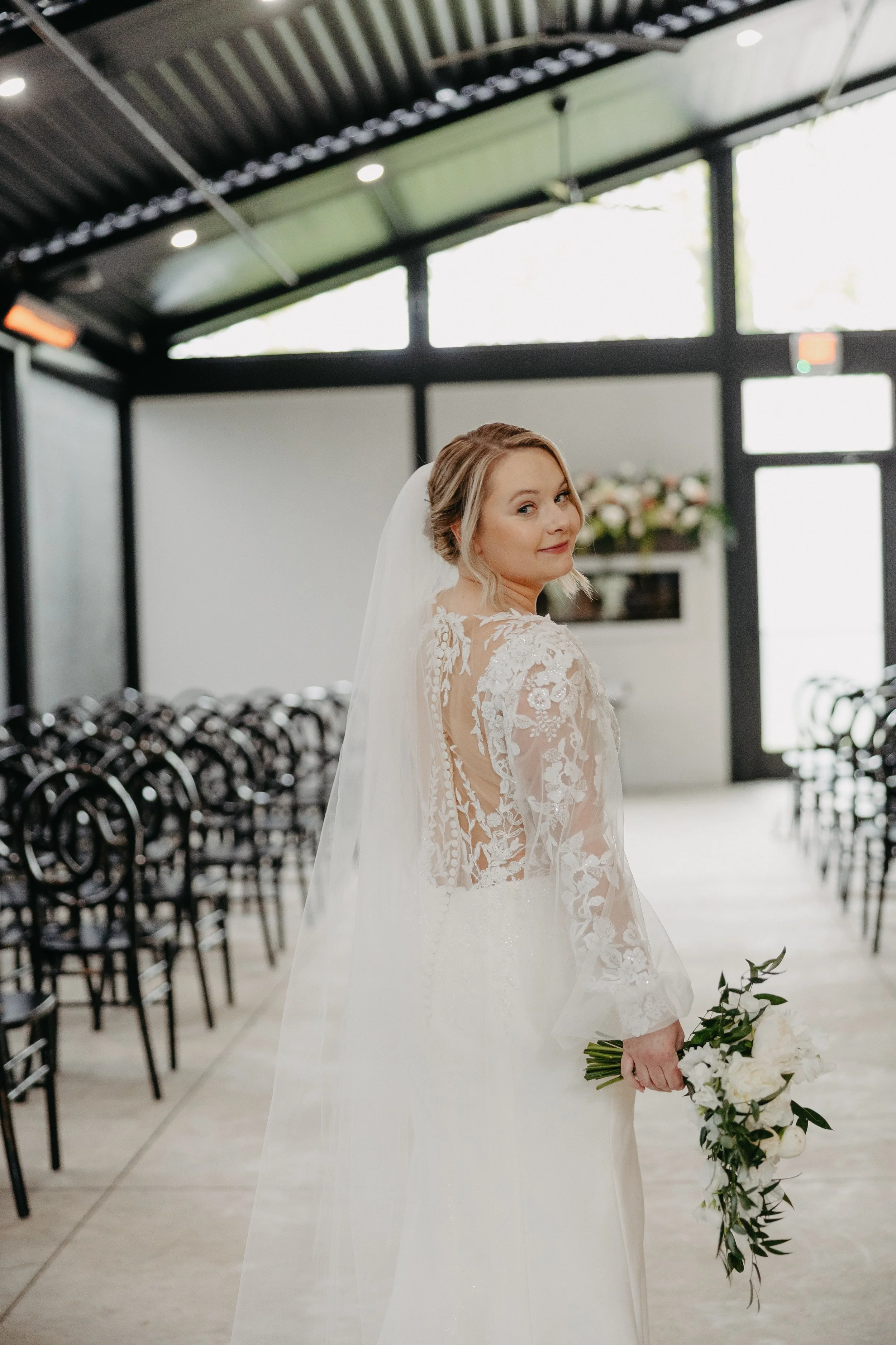 A bride in a white lace wedding dress with a veil holding a bouquet of white flowers and greenery inside a modern venue with black chairs arranged in rows and floral decorations in the background.