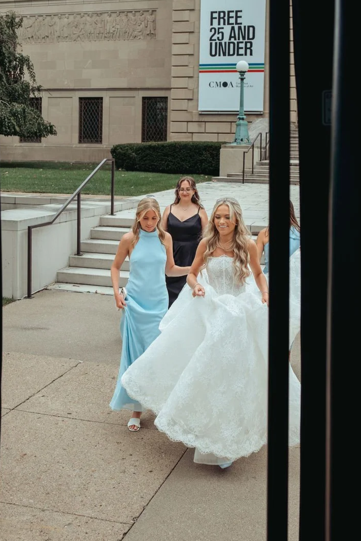 A bride in a white wedding gown walking outside with three bridesmaids in dresses, one in black, and the other in light blue. They are near steps in front of a building with a sign that says 'FREE 25 AND UNDER'.