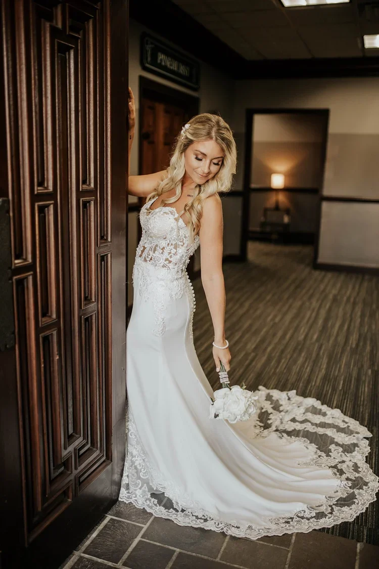 Bride in a white lace wedding dress holding a bouquet, leaning against a doorframe in a dimly lit indoor setting.