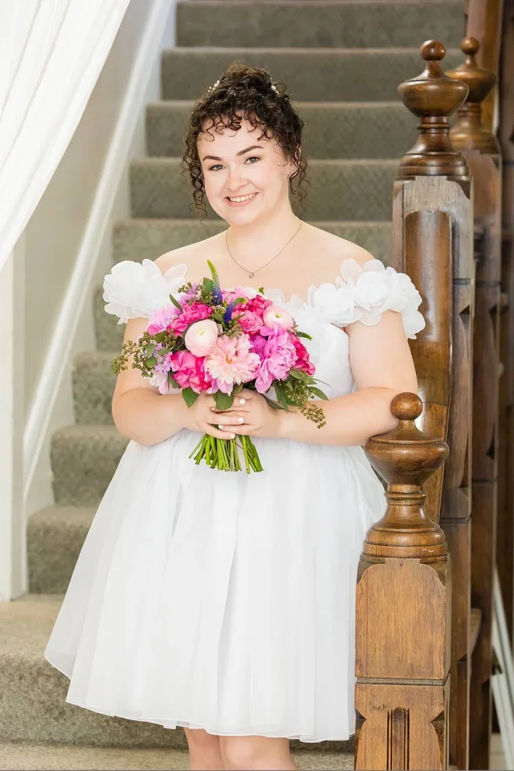 A young woman in a white dress holding a bouquet of pink and purple flowers, standing on a staircase with a wooden banister, smiling at the camera.