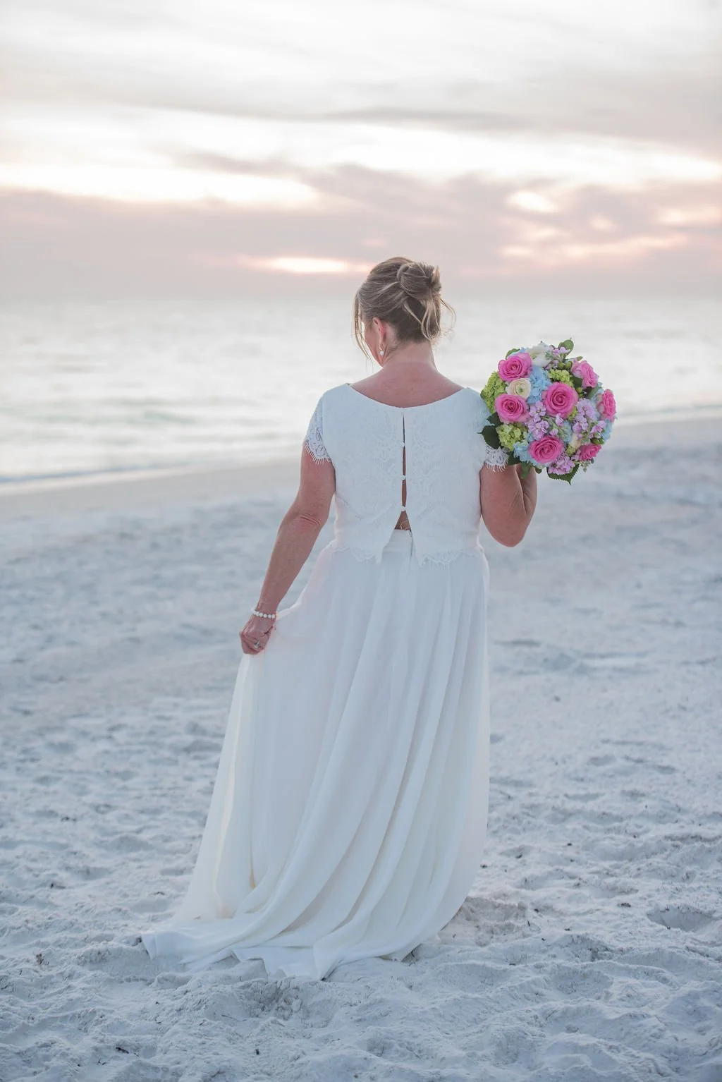 A woman in a white dress holding a colorful bouquet of flowers, standing on a sandy beach facing the ocean with a sunset sky in the background.