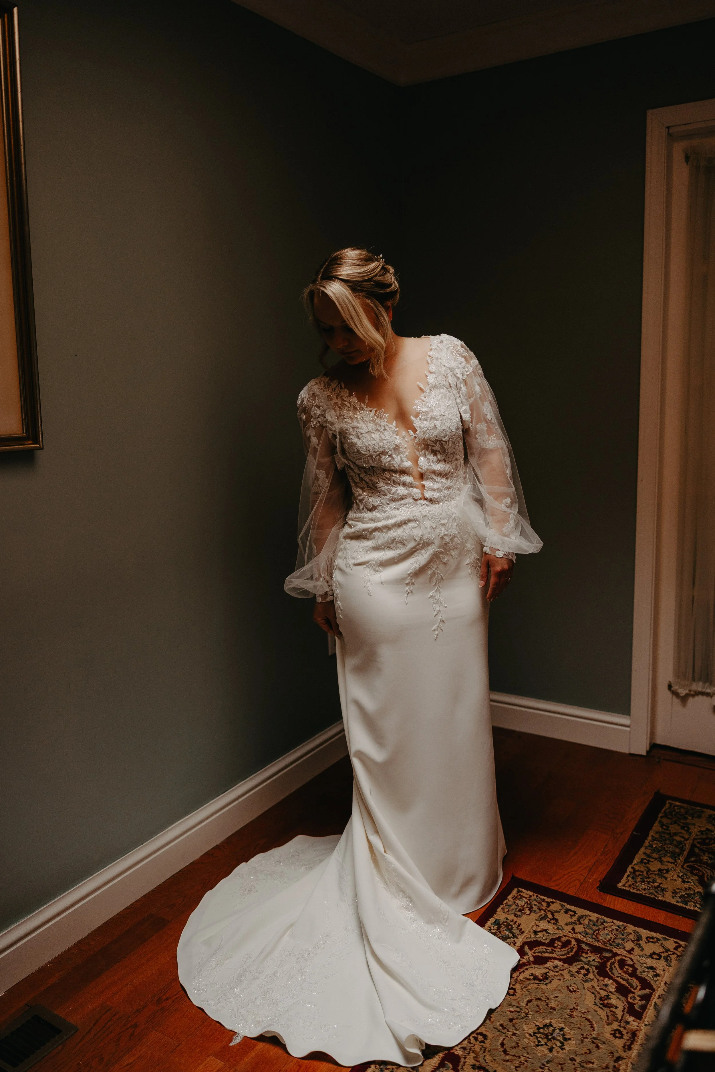 A woman dressed in an elegant wedding gown with lace details, standing in a dimly lit room with dark green walls, wooden floor, and patterned rug, looking down.