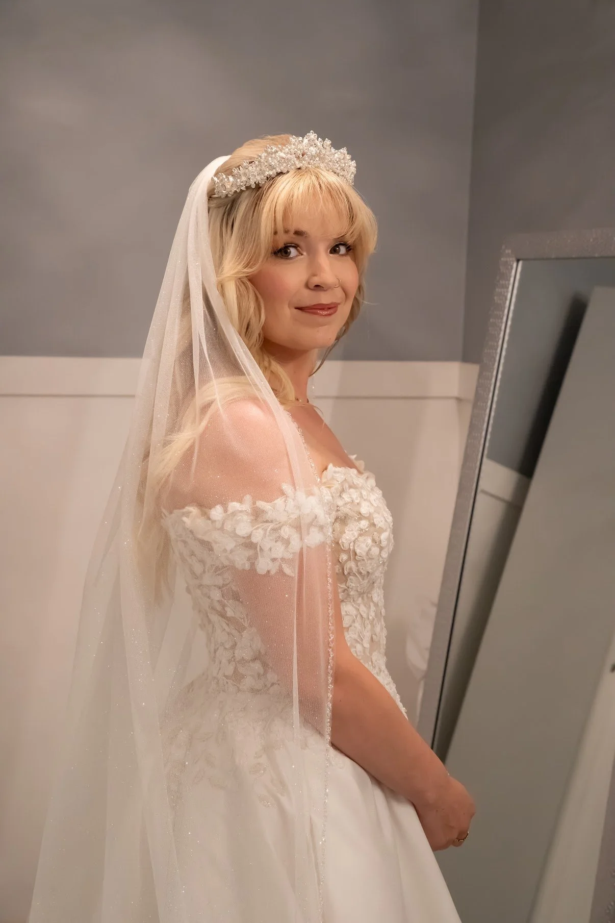 Young woman in a wedding dress with off-the-shoulder floral lace, wearing a pearl and crystal tiara, veil, and smiling softly, standing in front of a mirror.