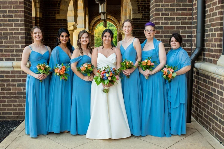 Group of seven women, one in a white wedding gown and six in matching blue bridesmaid dresses, standing outside and holding bouquets of flowers.
