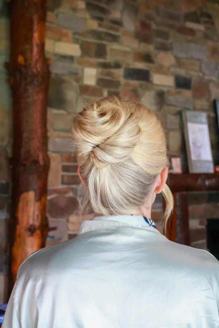 Back of a woman's head with elegant blonde hair styled in an updo, wearing a light-colored silk robe, with a brick wall and wooden beams in the background.