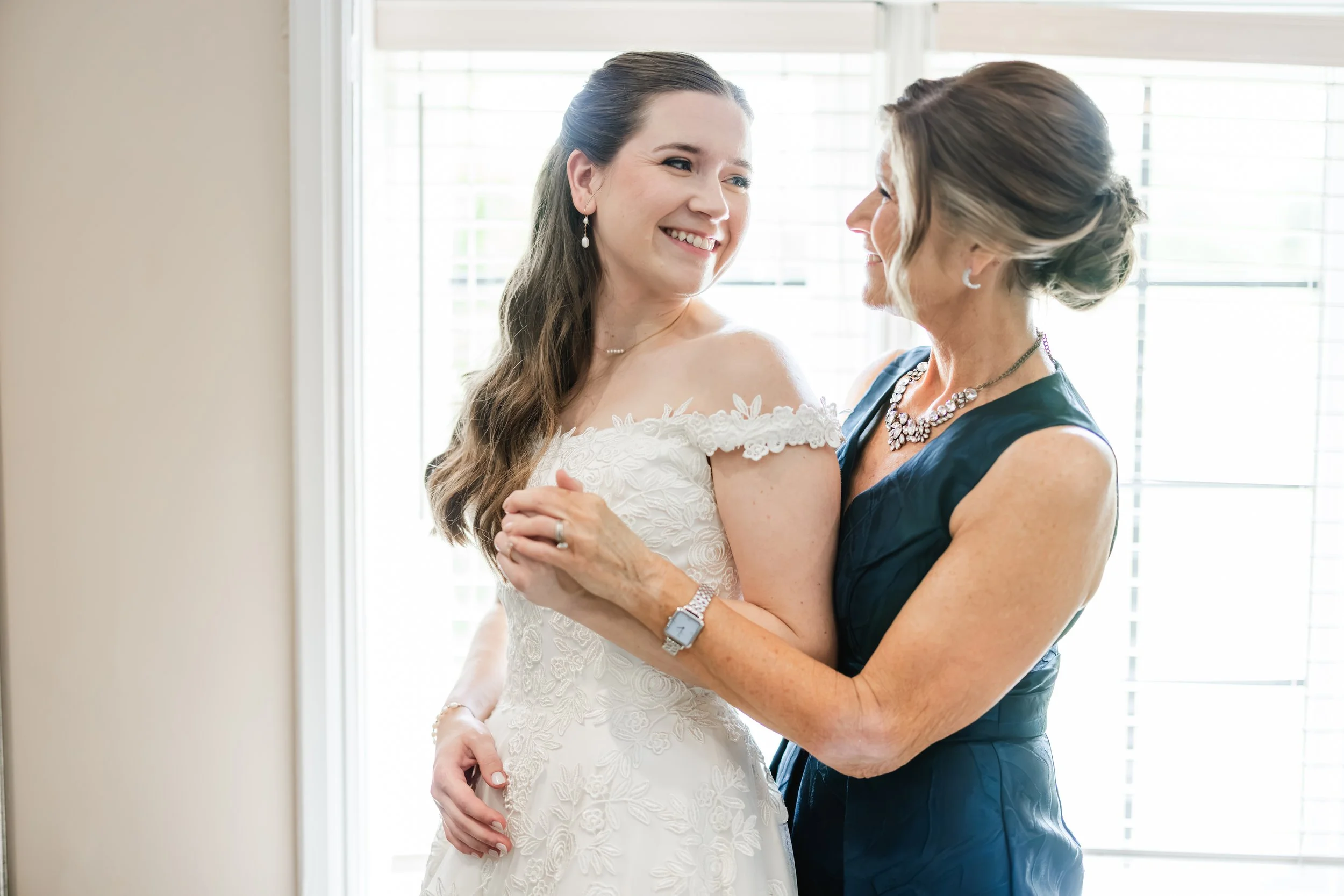 A young woman in a white wedding dress smiling and embracing an older woman in a dark blue dress, indoors with natural light coming through window blinds.