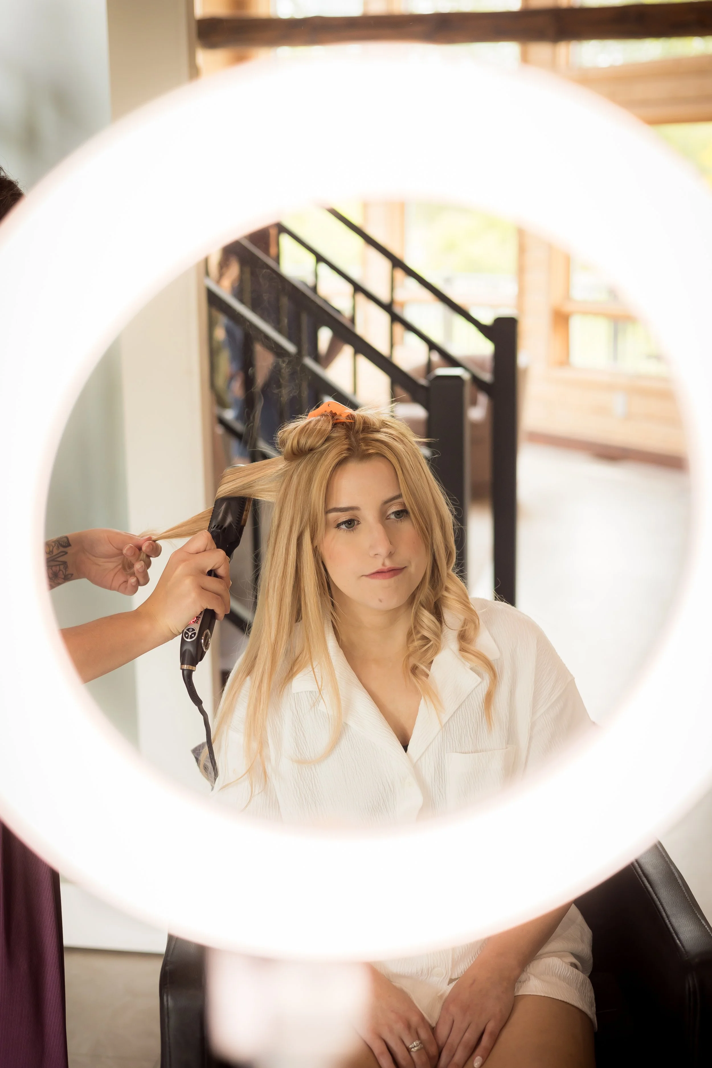 A woman with blonde hair sitting in a salon chair getting her hair blow-dried by a stylist. The photo is taken through a circular mirror or ring light that frames her face.