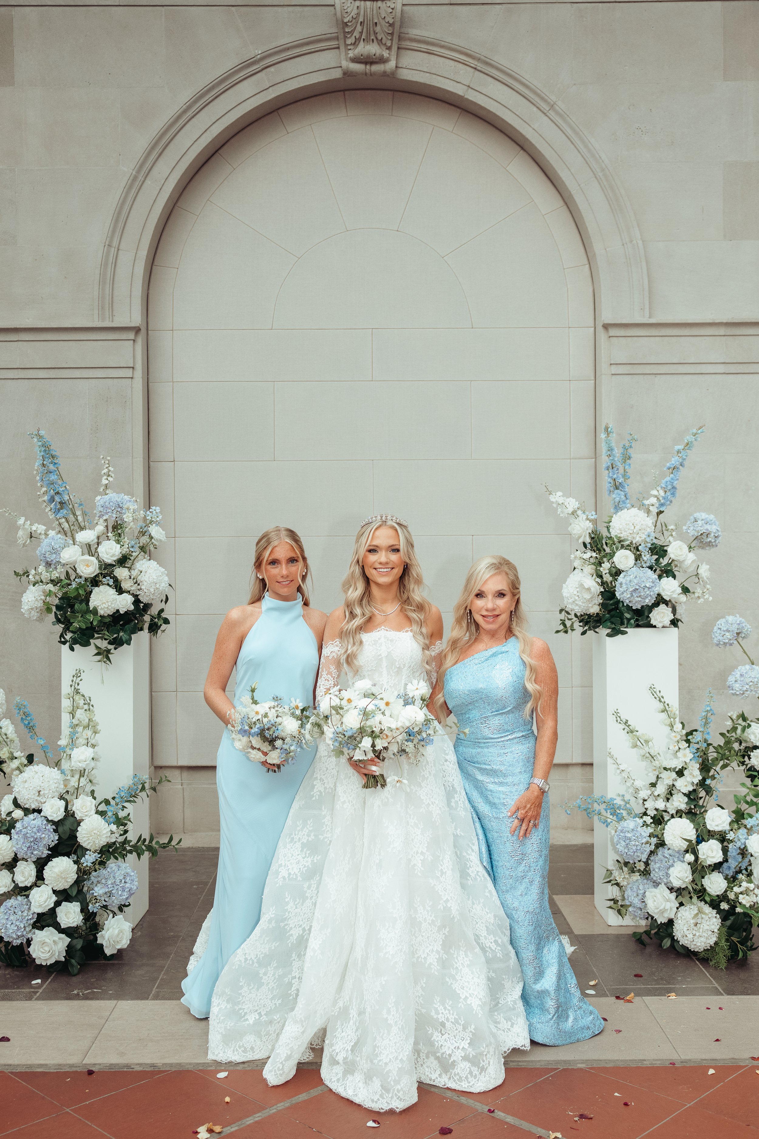 Three women in formal dresses standing in front of a light-colored wall with floral arrangements on either side. The woman in the center wears a white wedding gown with lace details and a tiara, holding a bouquet of white and pale pink flowers. The w