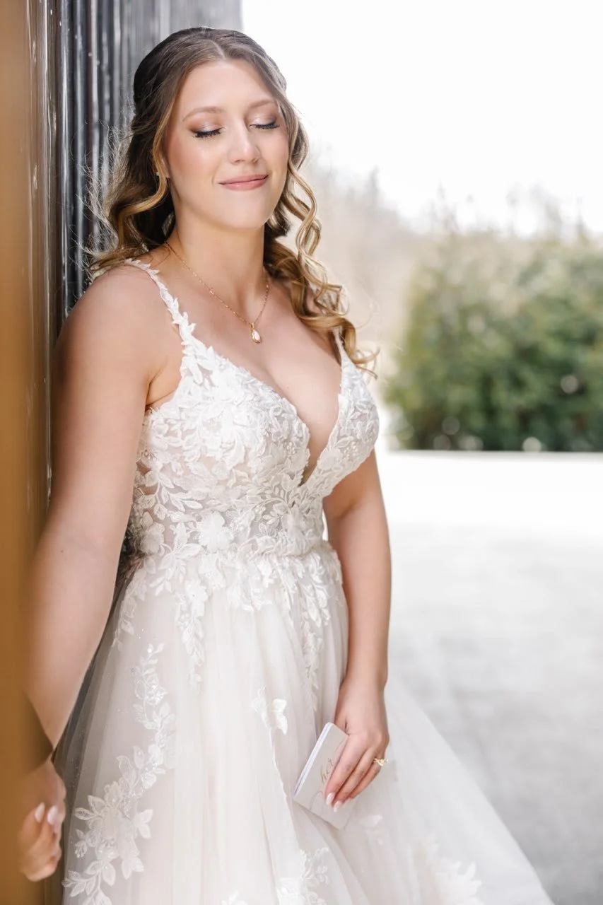 A bride in a white lace wedding gown with floral embroidery, standing indoors near a window with a soft smile and her eyes closed, holding a small white clutch.