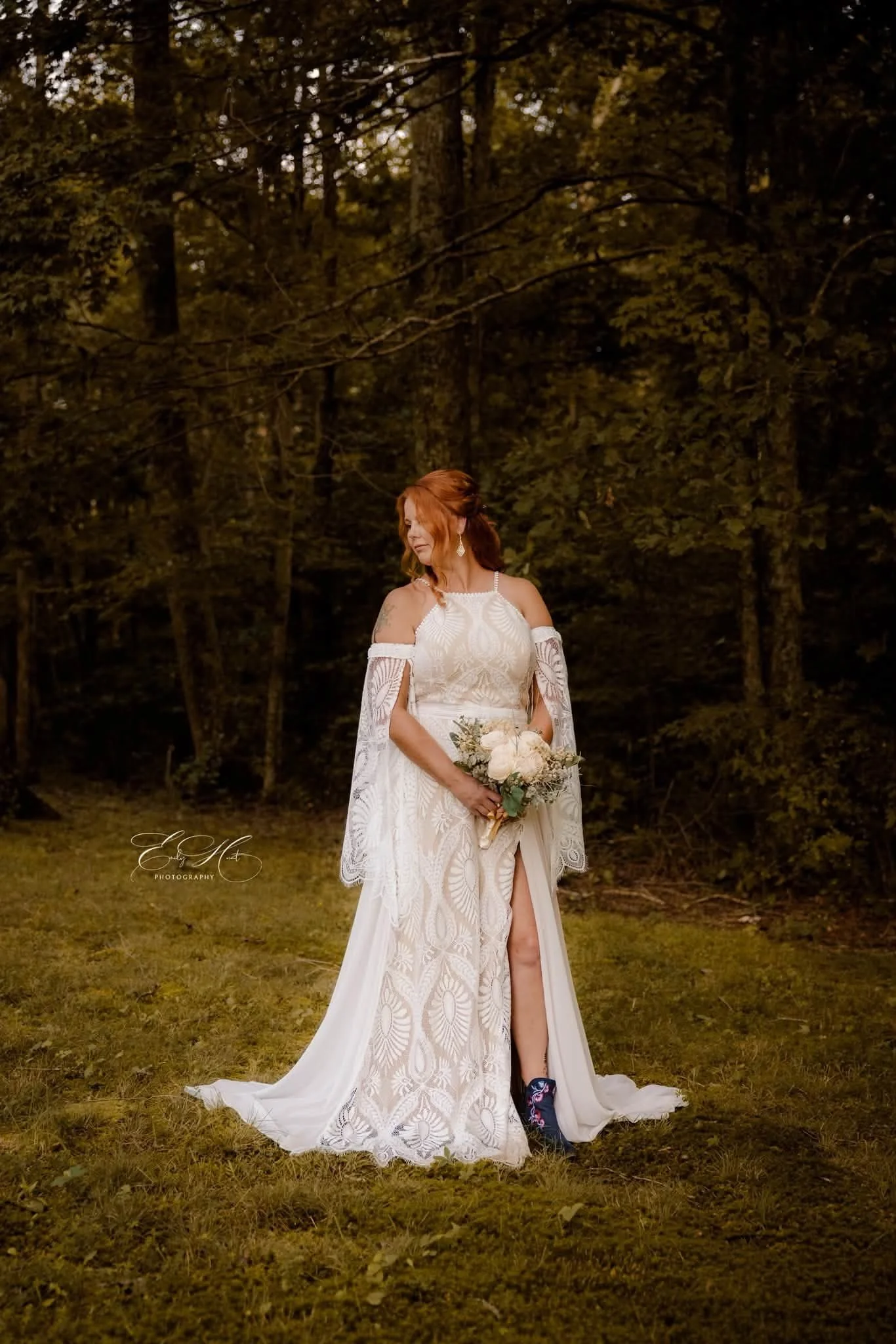 A woman in a white lace wedding dress holding a bouquet of white roses stands in a forested outdoor setting at dusk.