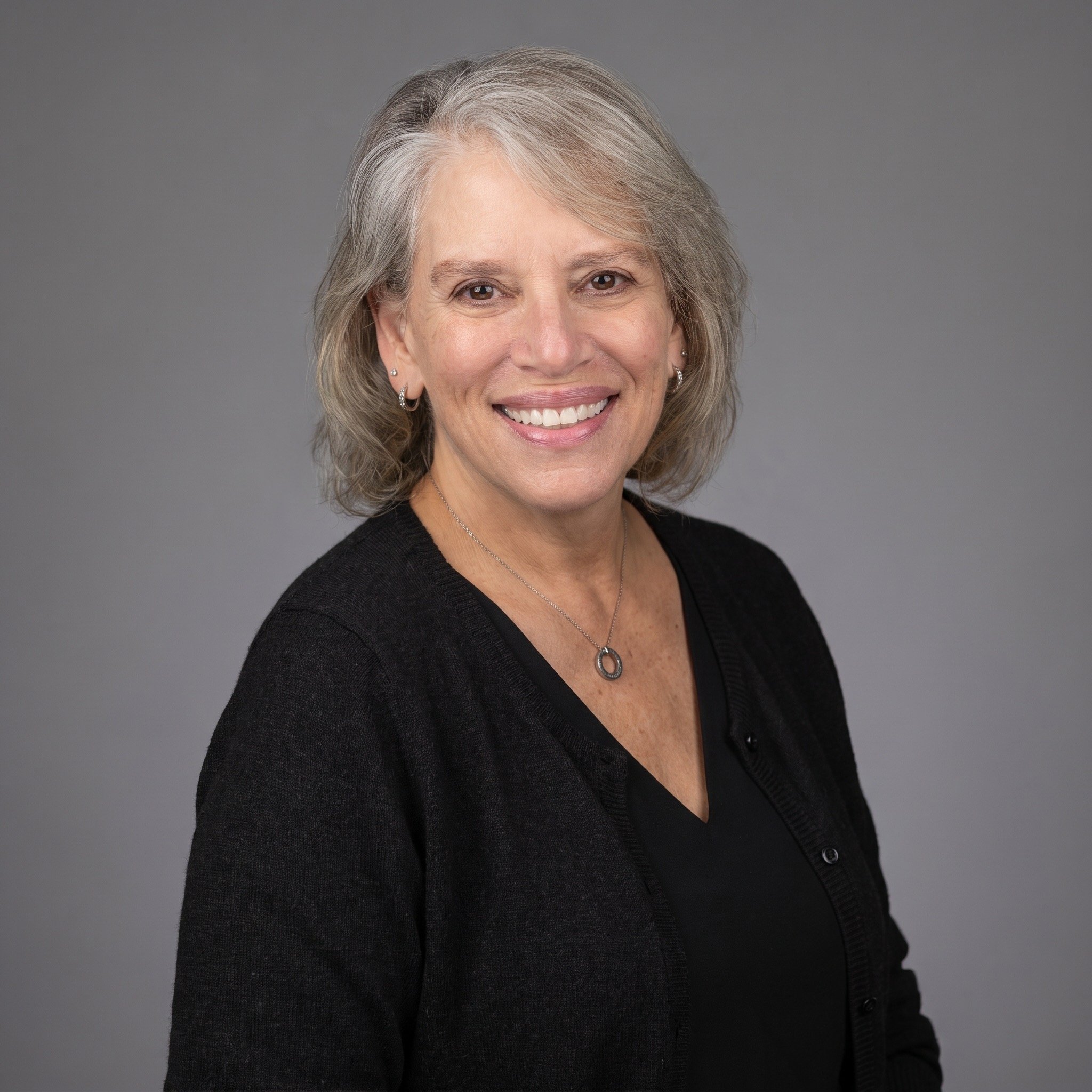 A headshot of a middle-aged woman with gray hair, smiling, set against a gray background. She is wearing earrings, a necklace, and a black top.