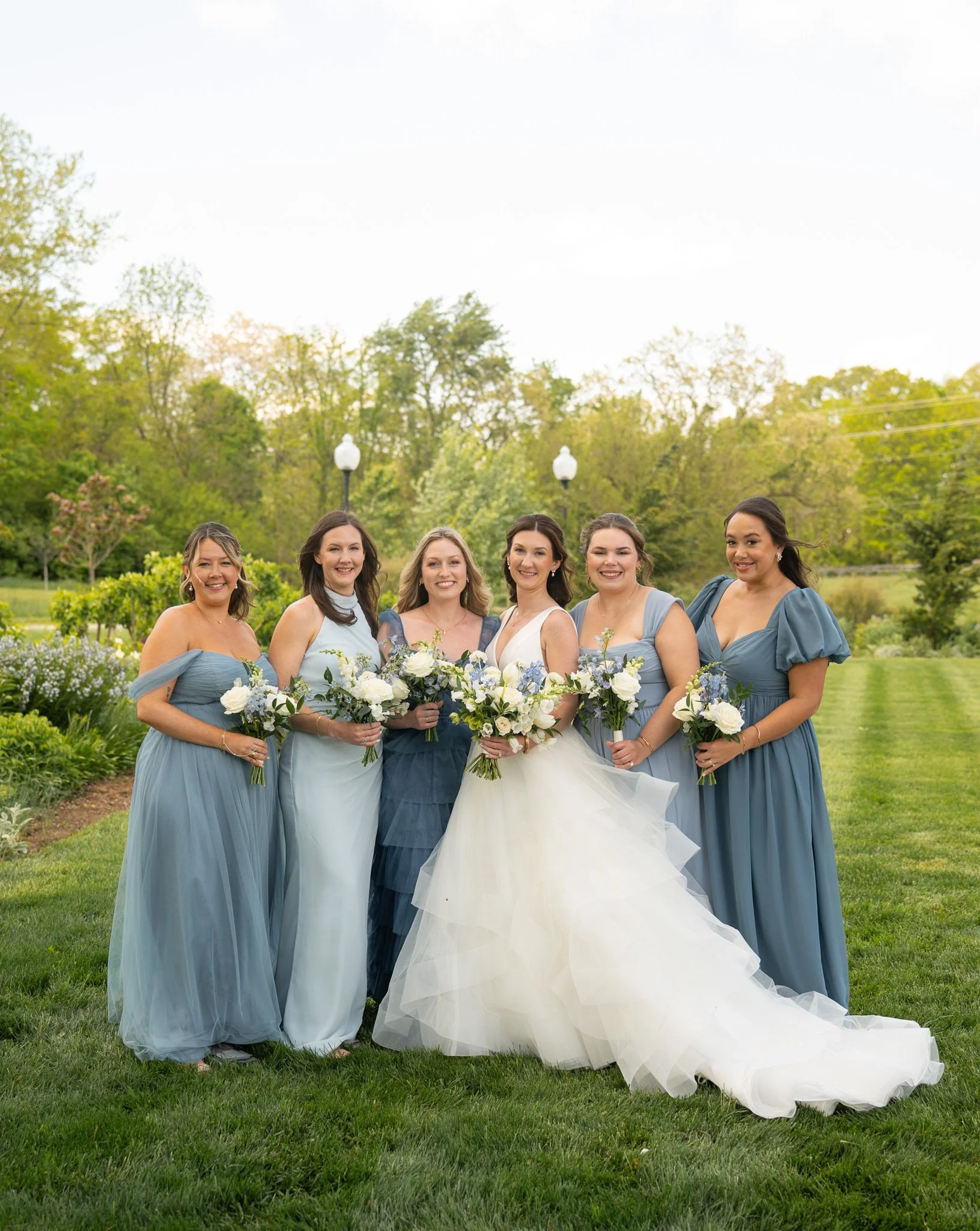 Group of seven women dressed in pastel blue and white bridesmaid dresses standing outdoors on a grassy lawn, holding bouquets of white and purple flowers, with trees and lamp posts in the background.