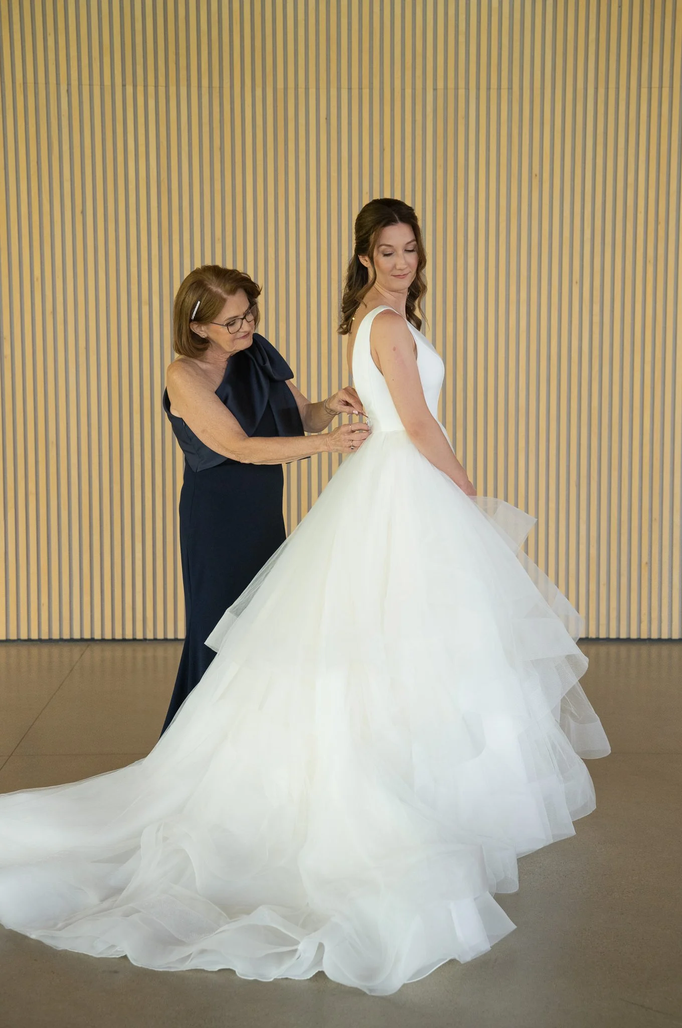 A bride in a white wedding dress and a woman in a navy dress adjusting the dress, standing against a wooden wall.