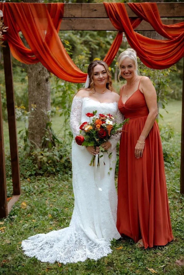 A bride in a white lace wedding dress holding a bouquet of red, orange, and white flowers stands next to a woman in a rust-colored dress. They are outdoors under a wooden arch decorated with orange drapes, surrounded by green trees and grass.