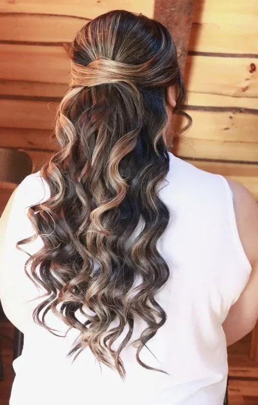 Back view of a woman with long, wavy hair styled with loose curls and a twisted half-up hairstyle, wearing a white sleeveless top, in front of a wooden wall background.