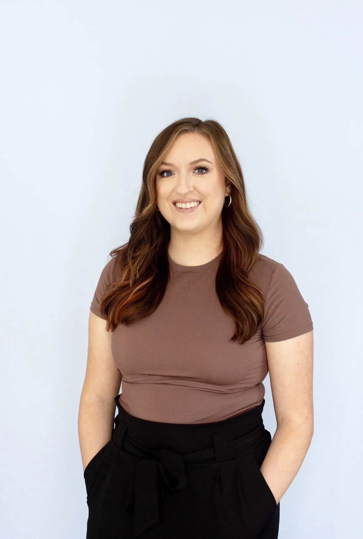 A young woman with long, wavy brown hair, blue eyes, and fair skin, smiling and wearing a brown T-shirt and black high-waisted pants with a bow tied at the waist, standing against a light blue background.