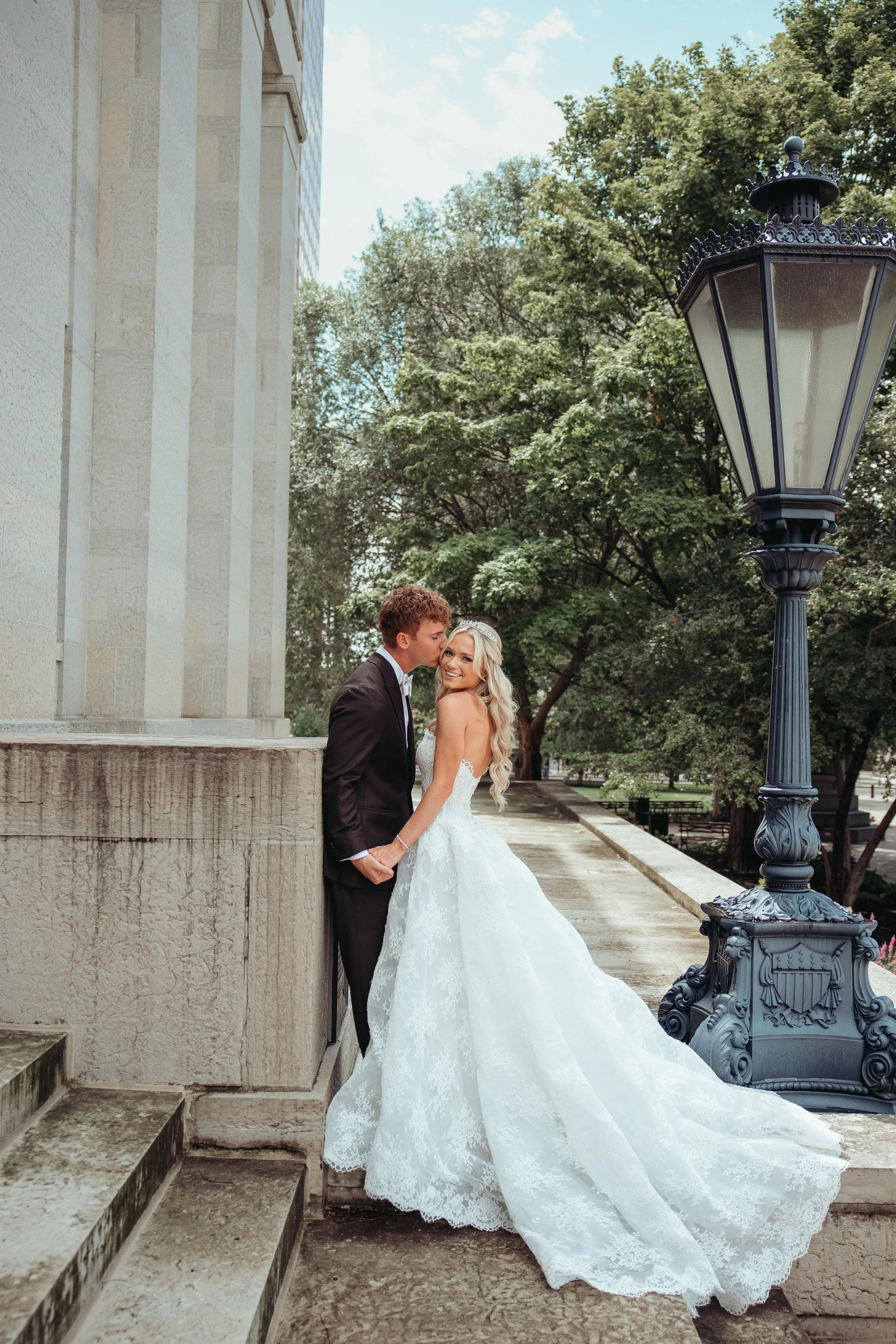 A newlywed couple standing outdoors near a stone building and a decorative street lamp, holding hands and sharing a moment of affection, with lush green trees in the background.