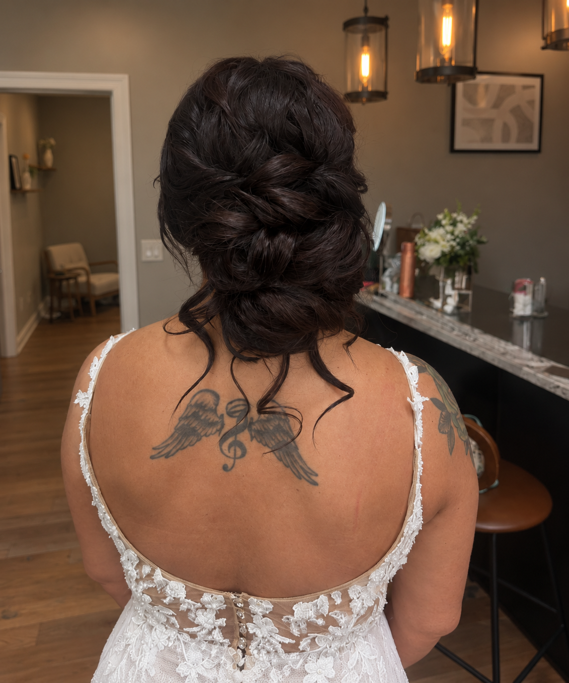 A woman with dark curly hair styled in an updo, wearing a white lace dress, shows a tattoo of angel wings and a musical staff on her upper back.