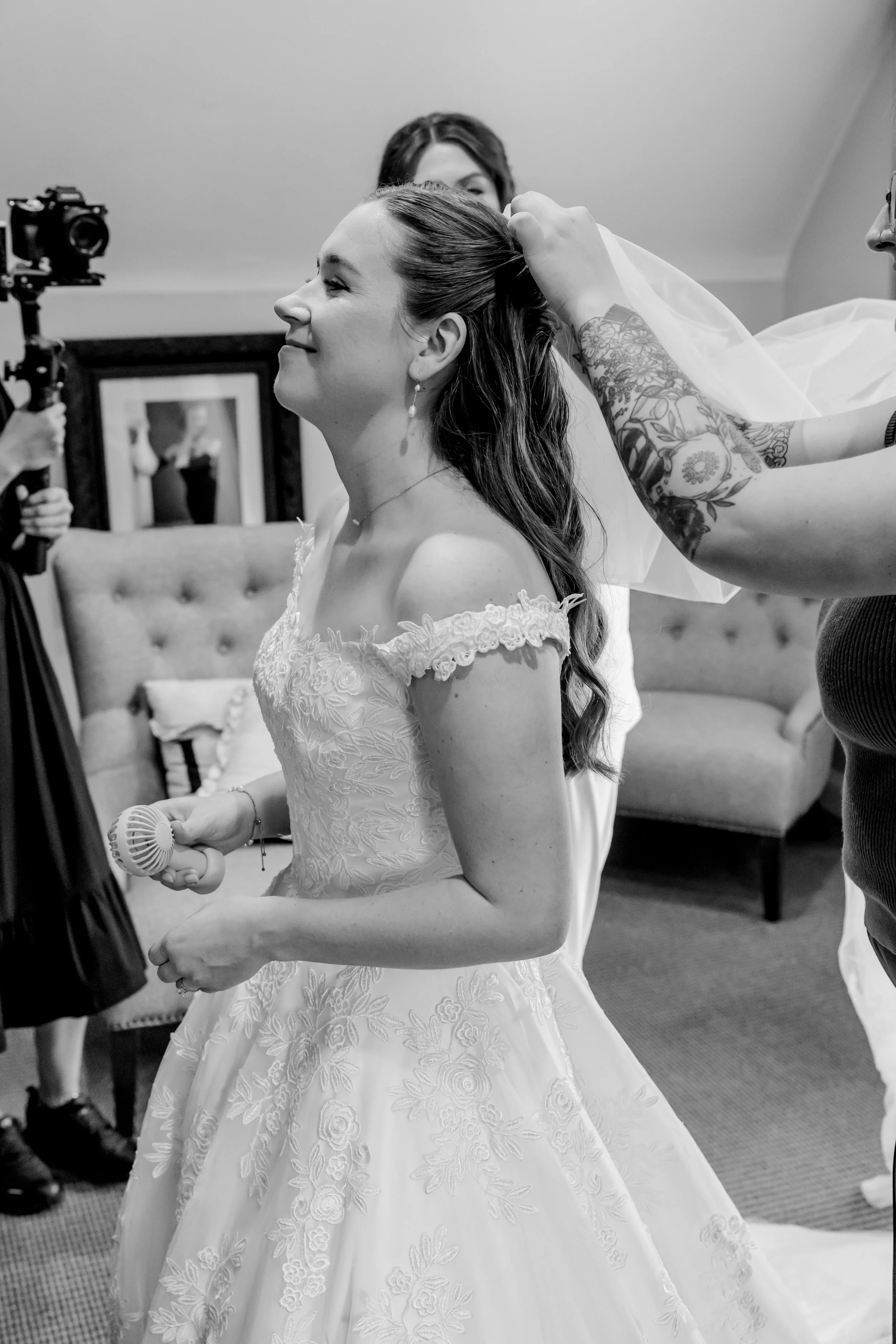 A bride with long hair in an off-shoulder wedding gown is smiling as someone adjusts her veil. A person with tattoos is helping her, and another woman is in the background, visible through a mirror. The scene is indoors, with a sofa and framed mirror