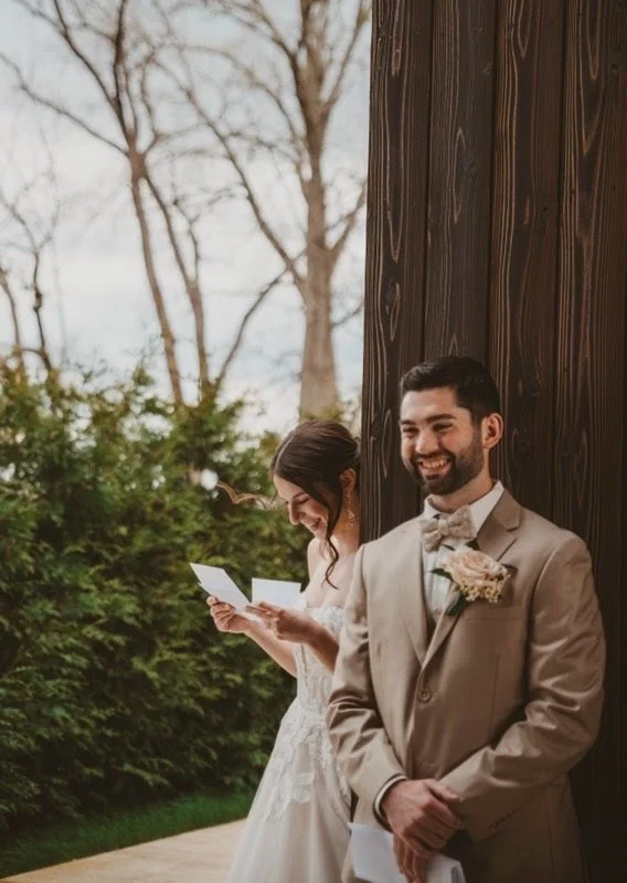 A bride and groom at their wedding, with the groom in a beige suit and the bride in a white wedding dress, standing outside near a dark wooden wall, with trees in the background. The groom is smiling with his hands clasped in front of him, while the 