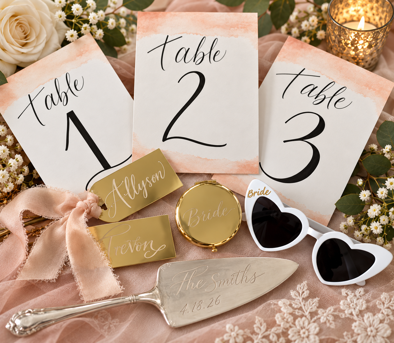 Wedding reception table markers and accessories, including table number cards labeled 1, 2, and 3, gold and white name tags for Allyson and Trevon, a gold mirror with 'Bride' engraved, heart-shaped sunglasses, a silver cake server engraved with 'The Smiths 4.18.26,' surrounded by flowers and a lit candle.