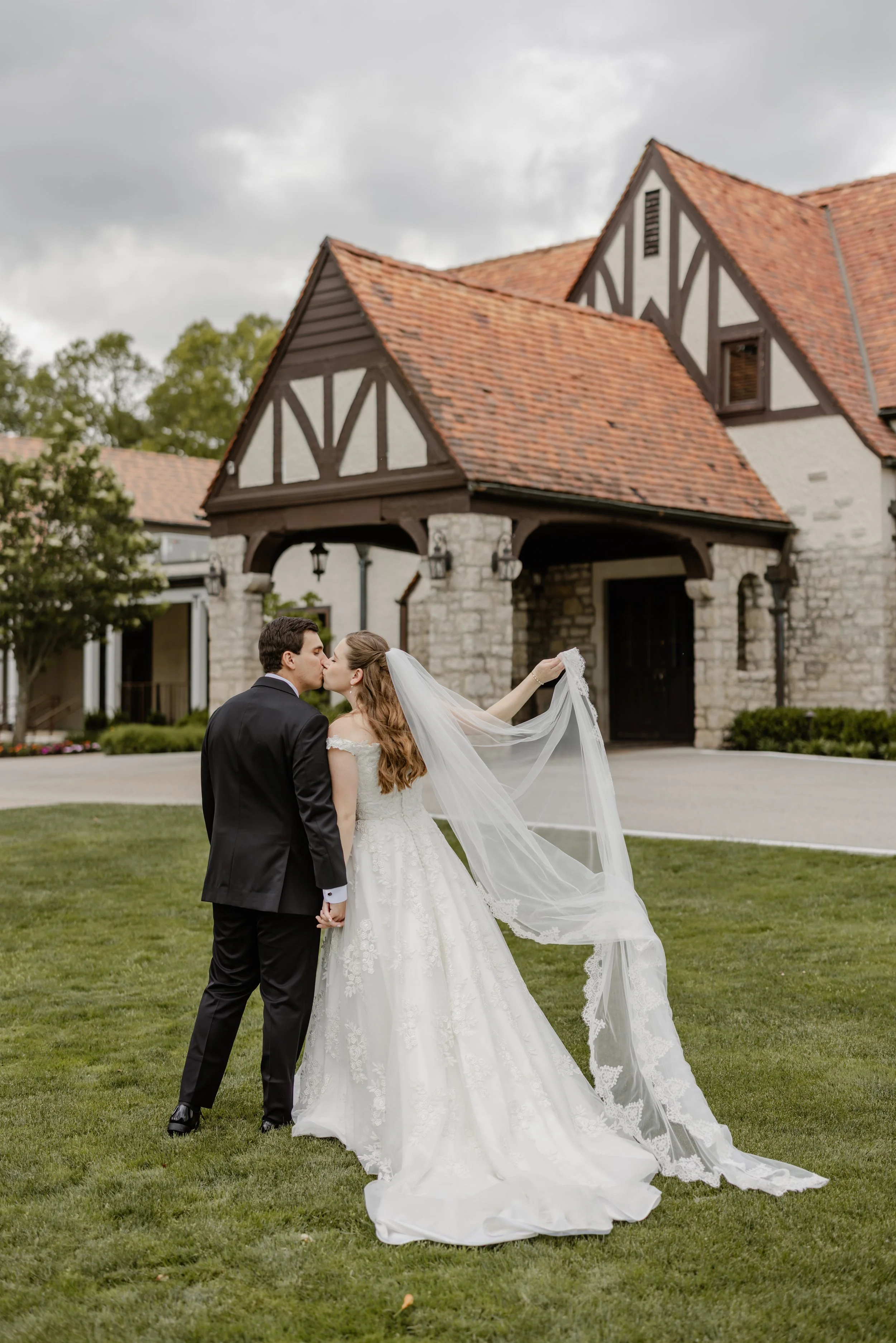 A bride and groom sharing a kiss outdoors in front of a rustic stone and wood building on a cloudy day.