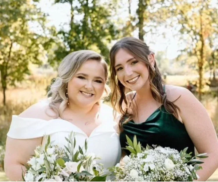 Two women at a wedding holding bouquets, smiling outdoors with fall foliage in the background.