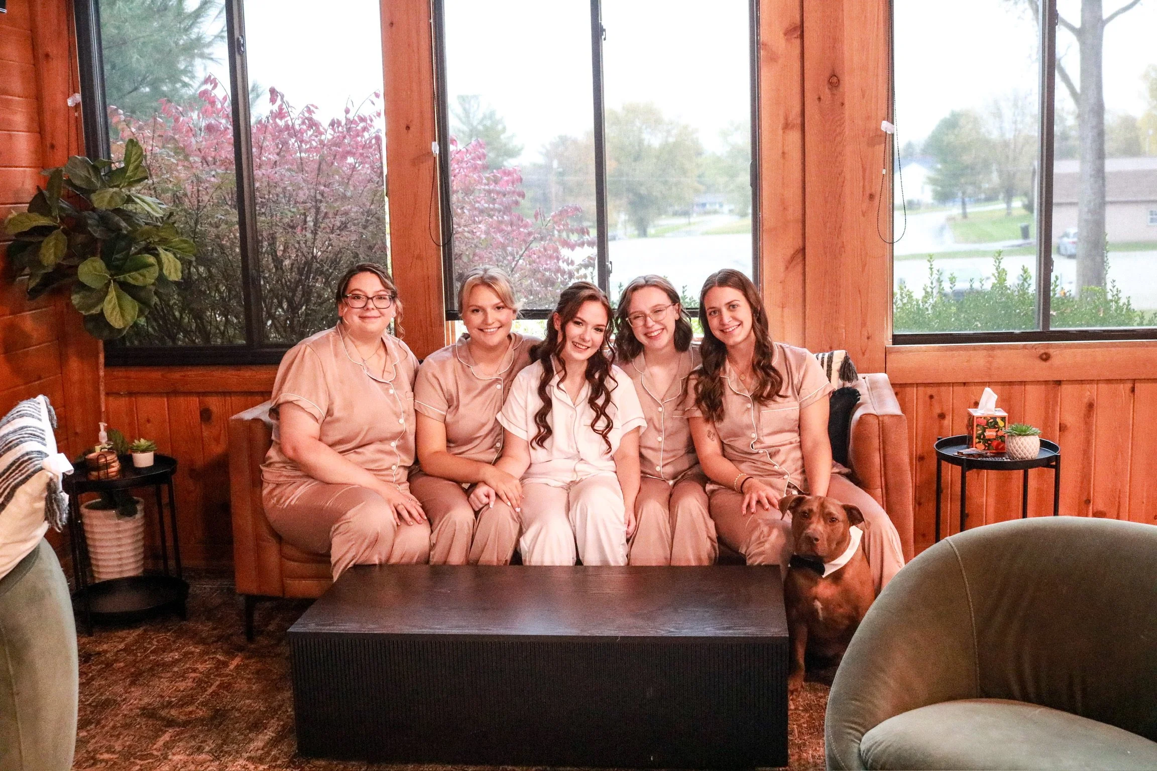 A group of five women, dressed in matching beige pajamas, sitting on a beige sofa in a room with wooden walls and large windows showing an outdoor scene with trees and houses. There is a dog sitting in front of the woman on the right.