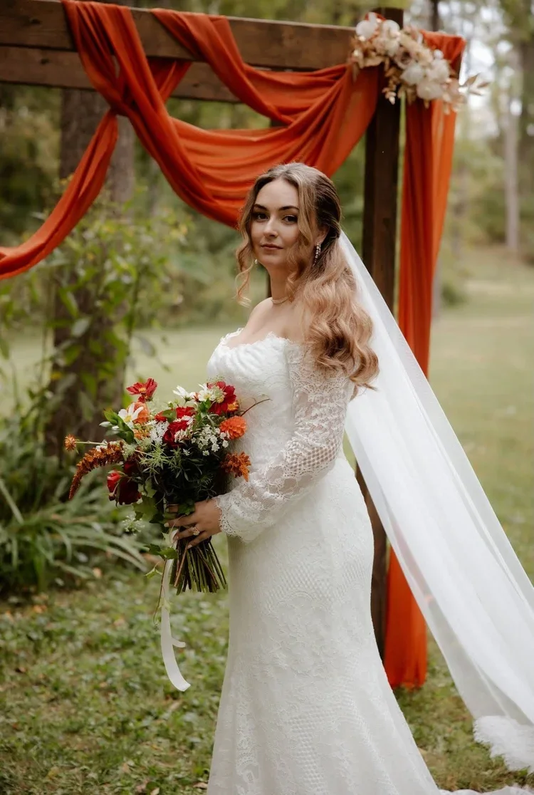 Bride in a white lace wedding dress holding a bouquet of red, orange, and white flowers outdoors with a wooden arch draped in orange fabric and floral decorations.