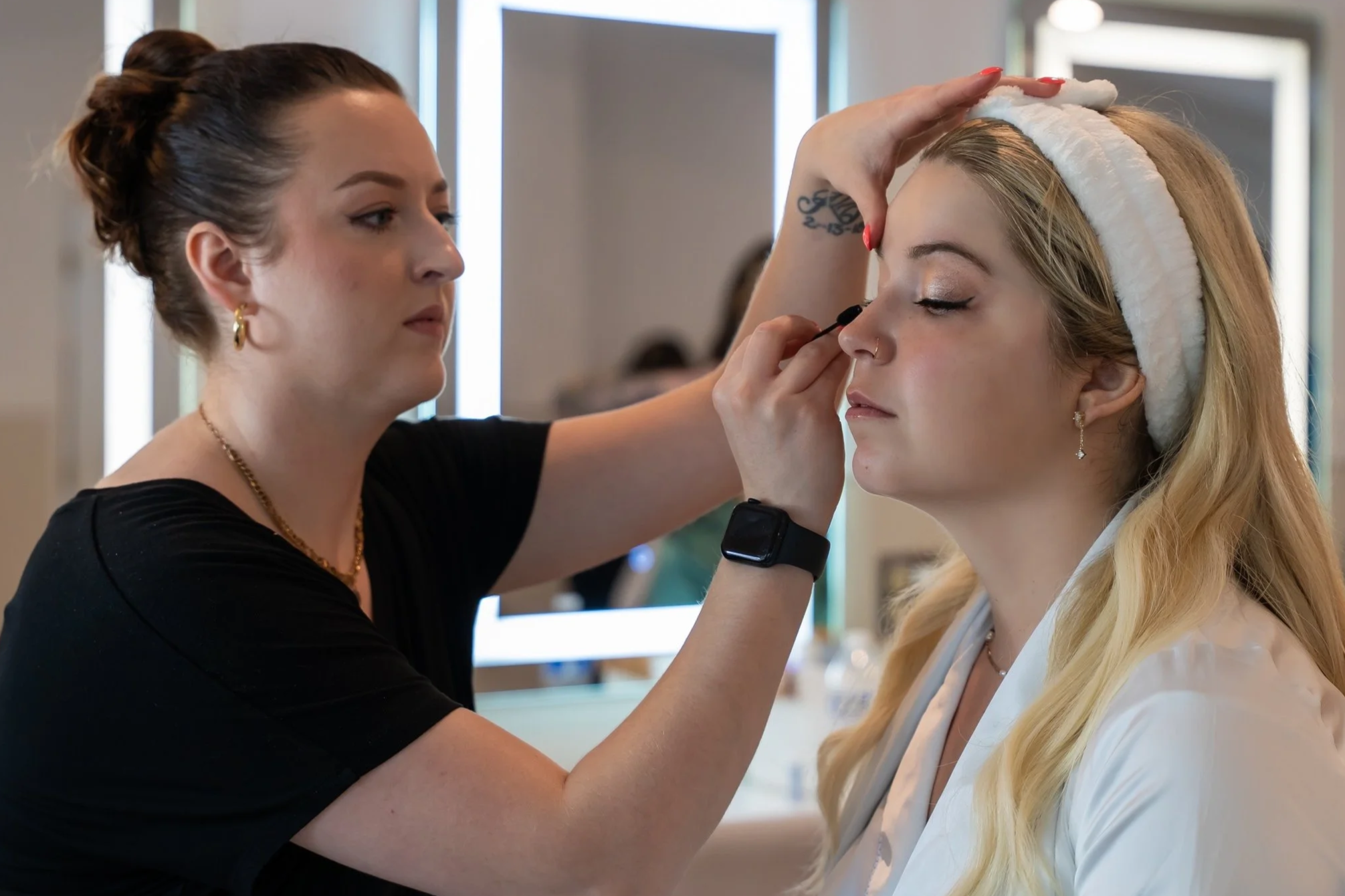 A makeup artist applies eye makeup to a woman with blonde hair wearing a white headband, in a brightly lit room.