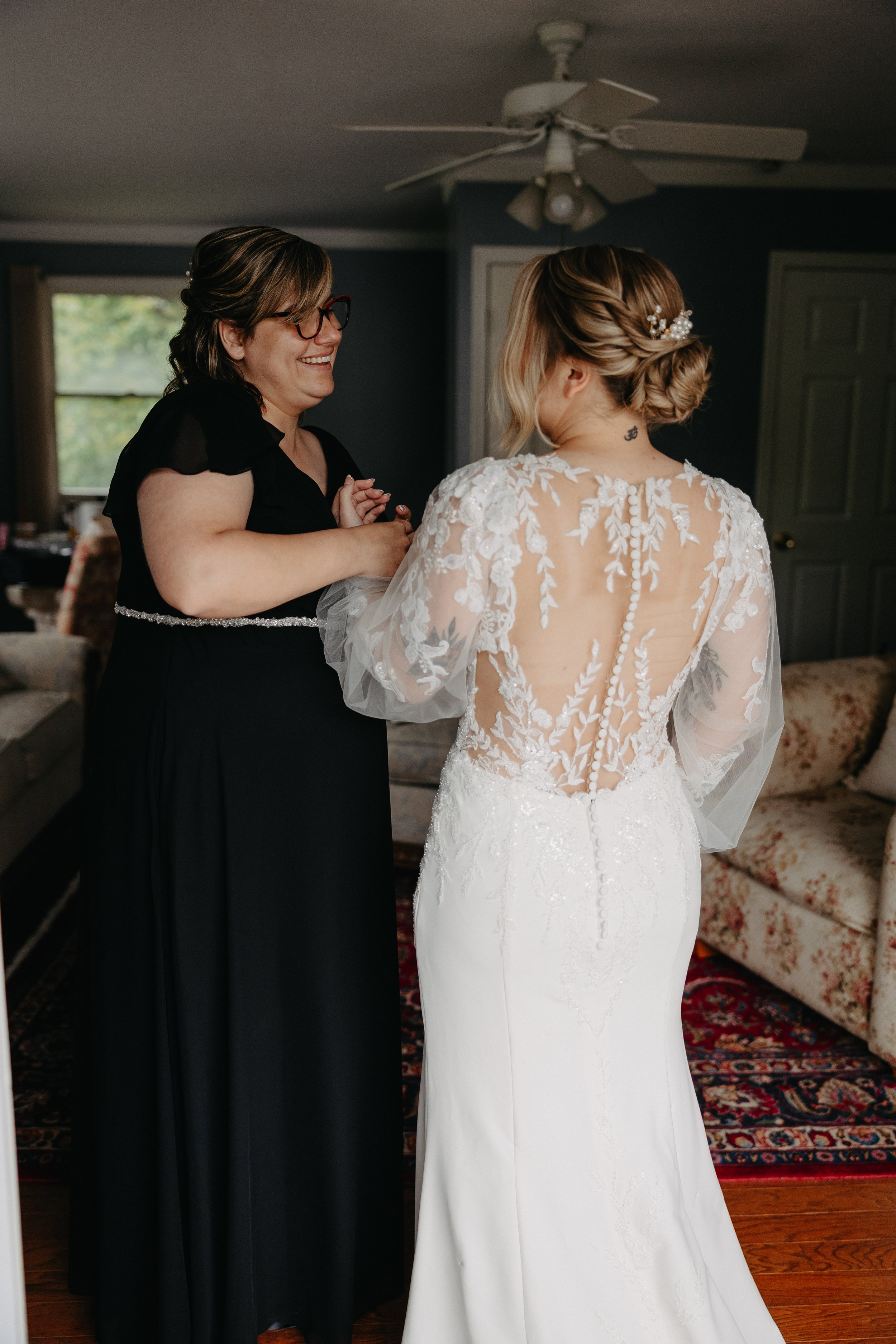 A woman dressed in a white wedding gown with lace detailing and a braid hairstyle with pearl hair accessories is facing a woman in a black dress with glasses, both are holding hands and smiling.