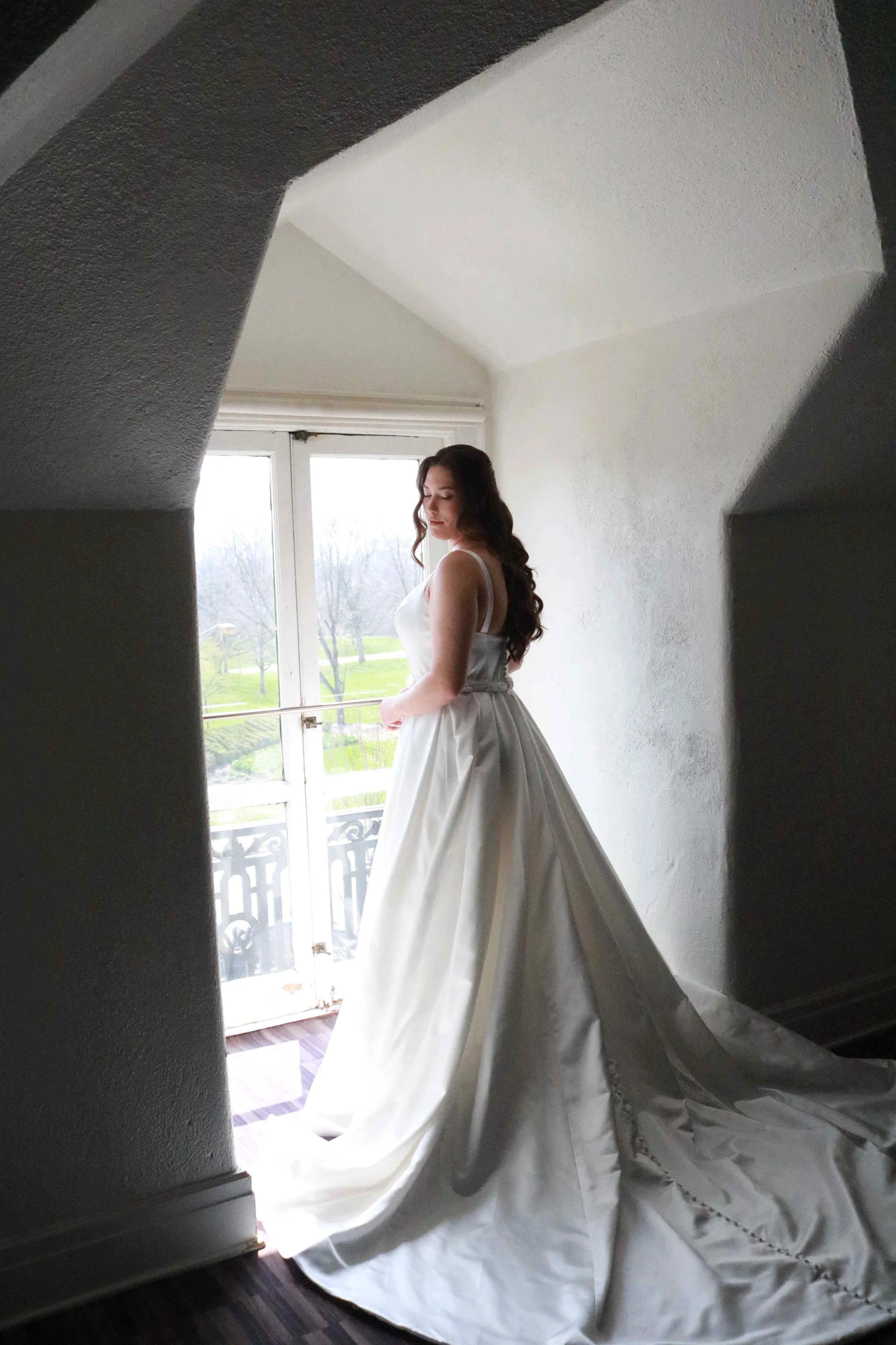 A woman in a white dress standing near a window, casting a soft light on her, with a white duvet or bedspread on the floor and draped over the window sill.