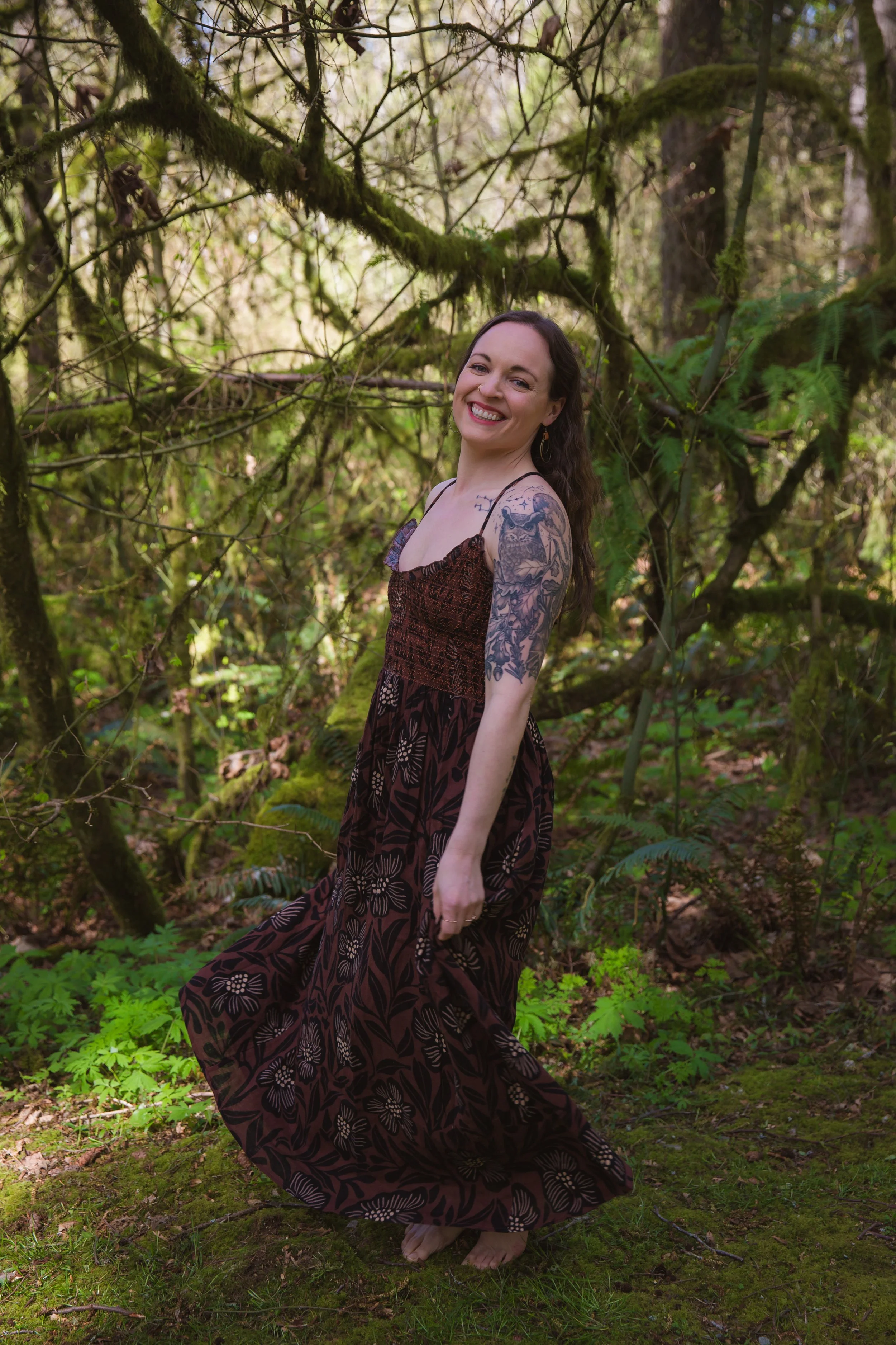 A woman with long dark hair smiling and standing barefoot in a forest with moss-covered trees and lush greenery, wearing a dark brown floral dress and tattoos on her arm.