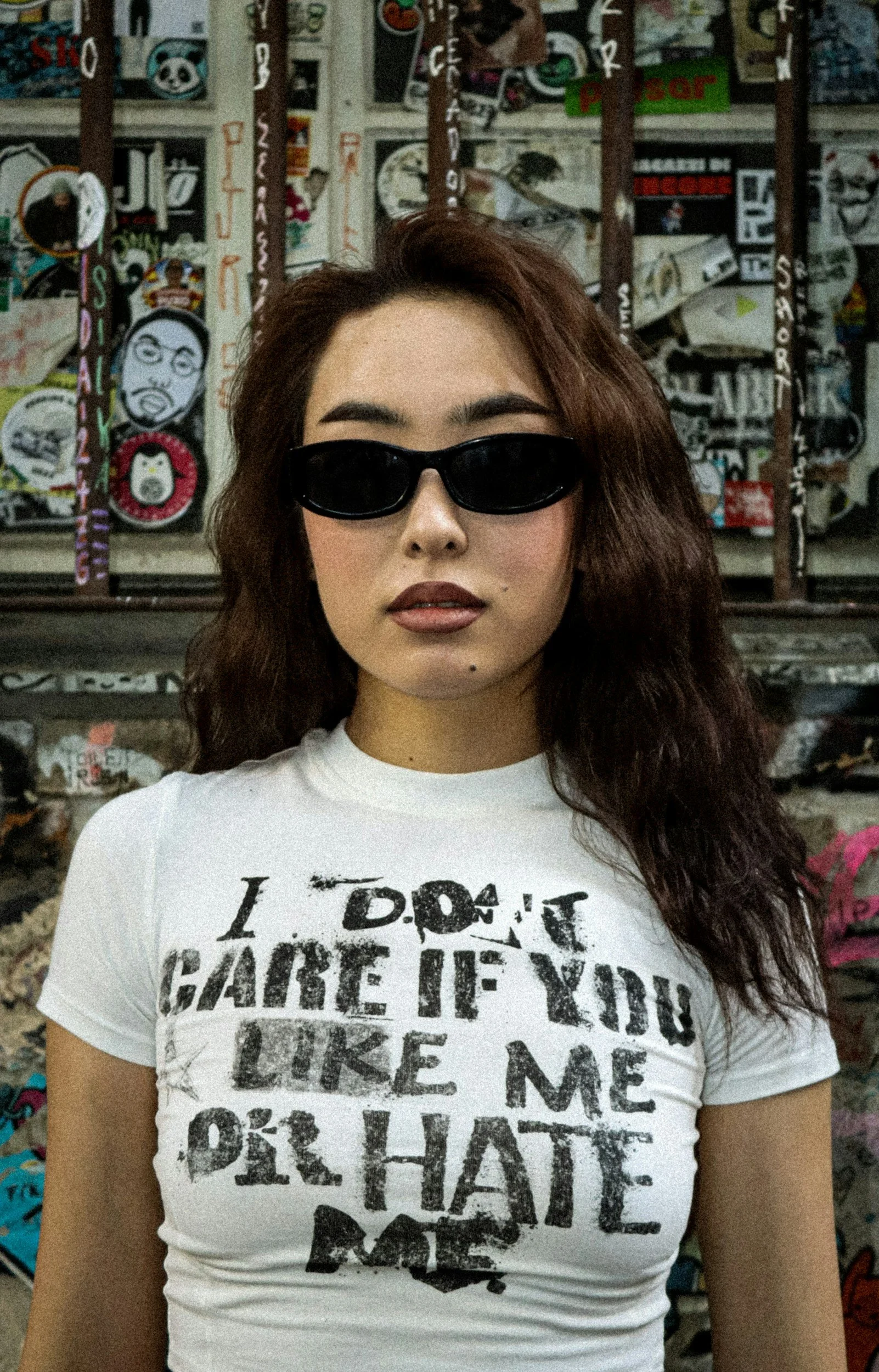 A woman with long wavy brown hair wearing black sunglasses and a white t-shirt with distressed black text standing in front of a graffiti-covered wall.