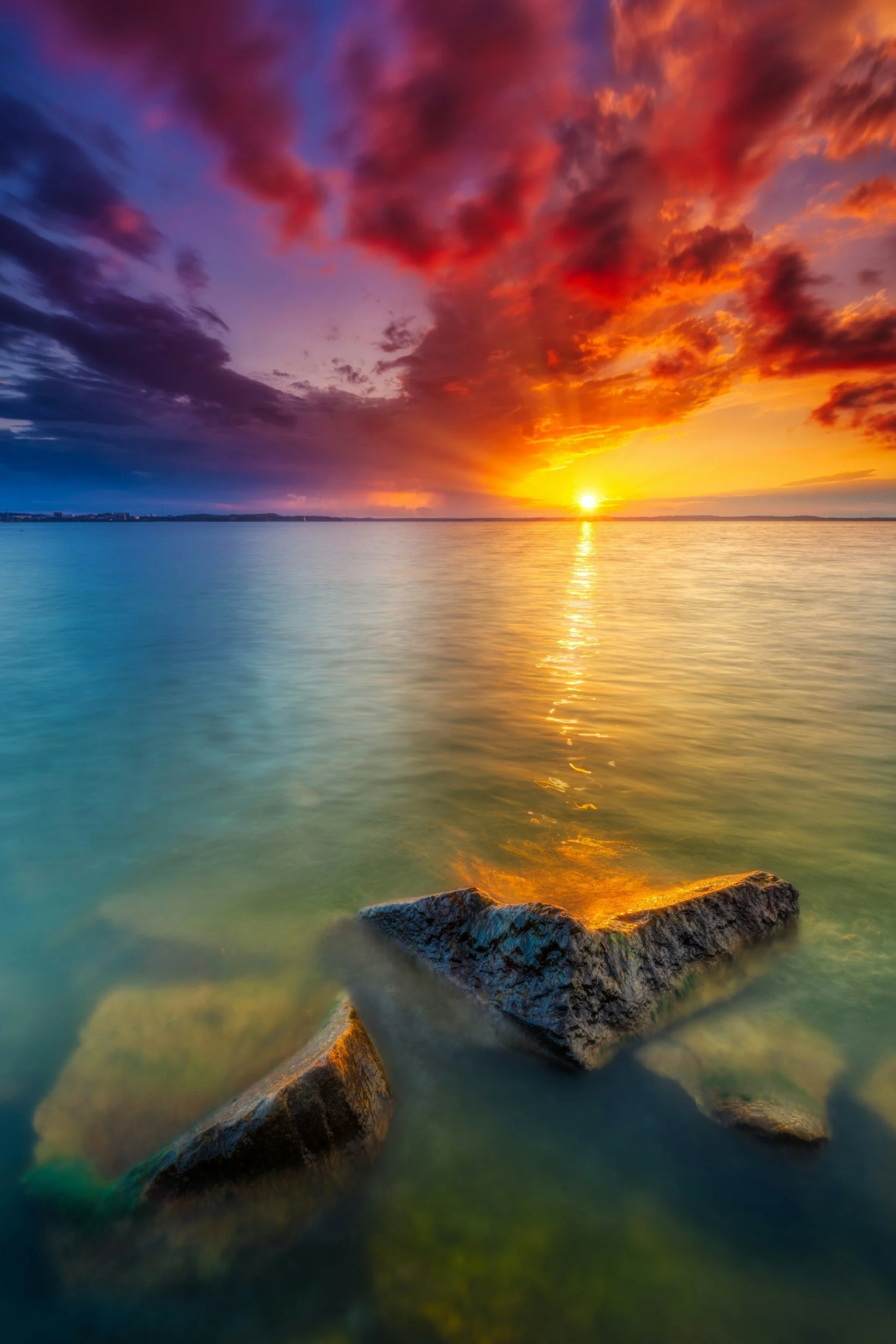A vibrant sunset over a calm body of water, with colorful clouds in shades of purple, pink, and orange, and rocks visible in the foreground.