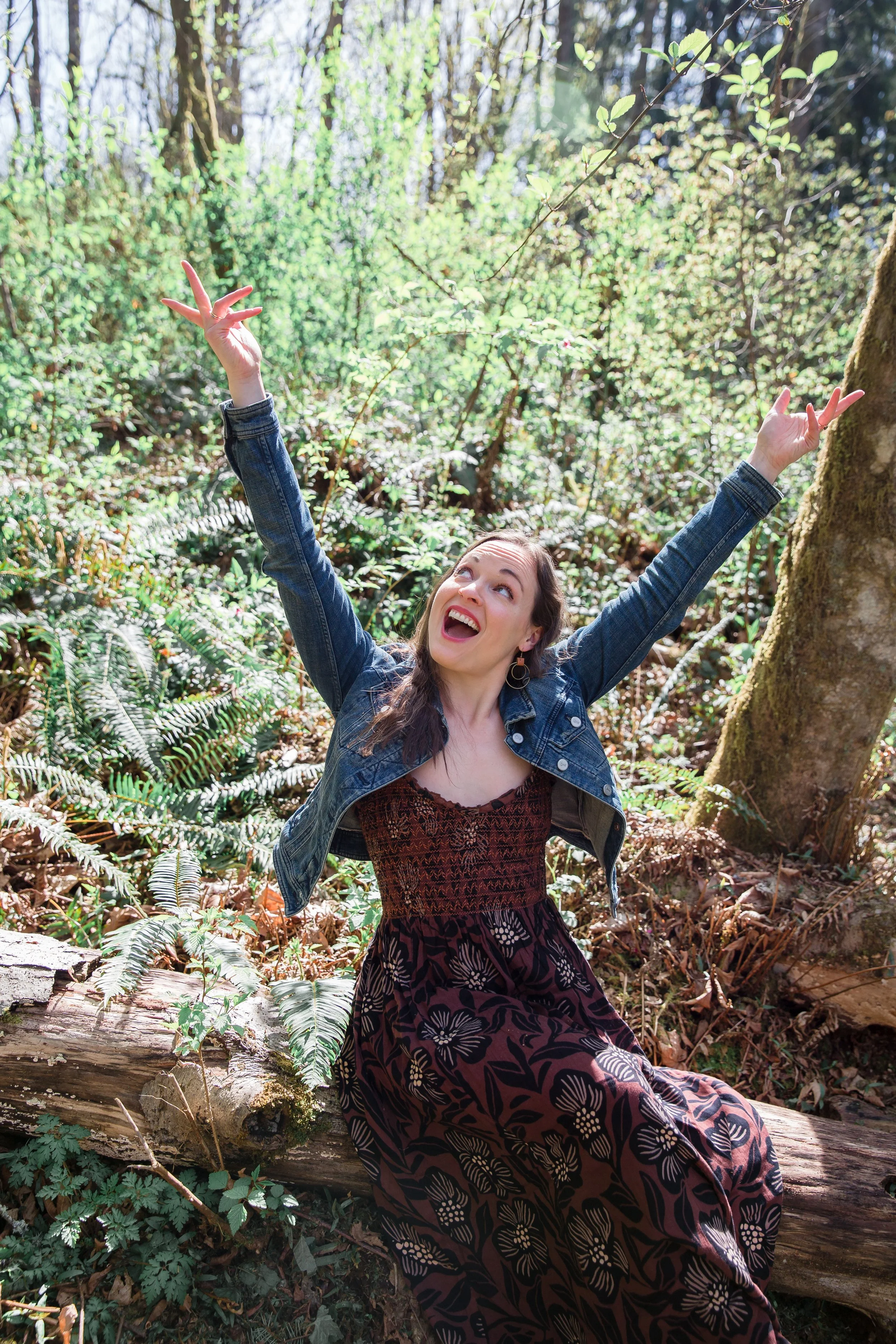A woman in a denim jacket and patterned dress sitting on a log in a lush forest, smiling and raising her arms joyfully.