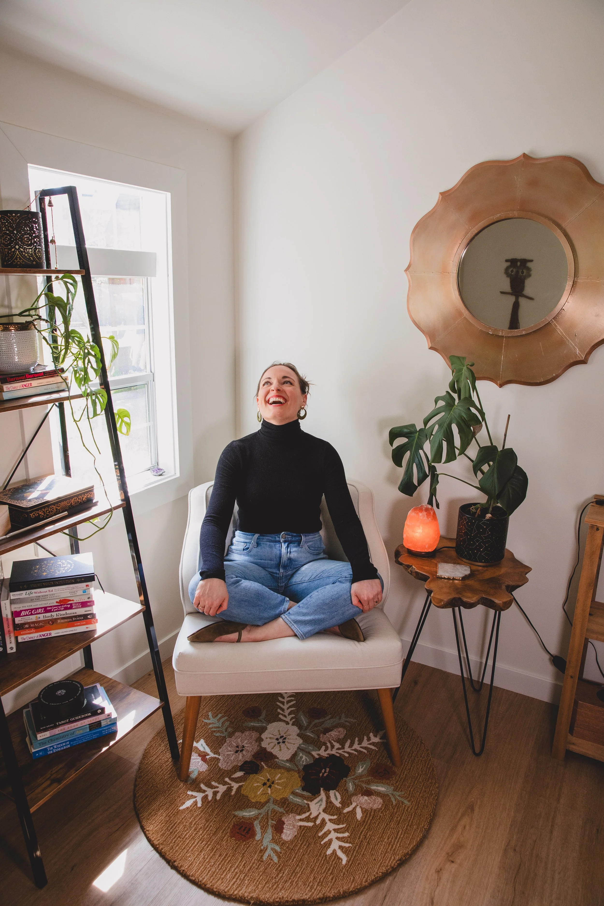 A woman with a black turtleneck and blue jeans sitting cross-legged on a white armchair in a cozy, well-lit room, smiling and looking upward. The room has a bookshelf filled with books, a wooden side table with a salt lamp, a large mirror with a decorative frame, and a tall plant. The floor has a round floral rug.
