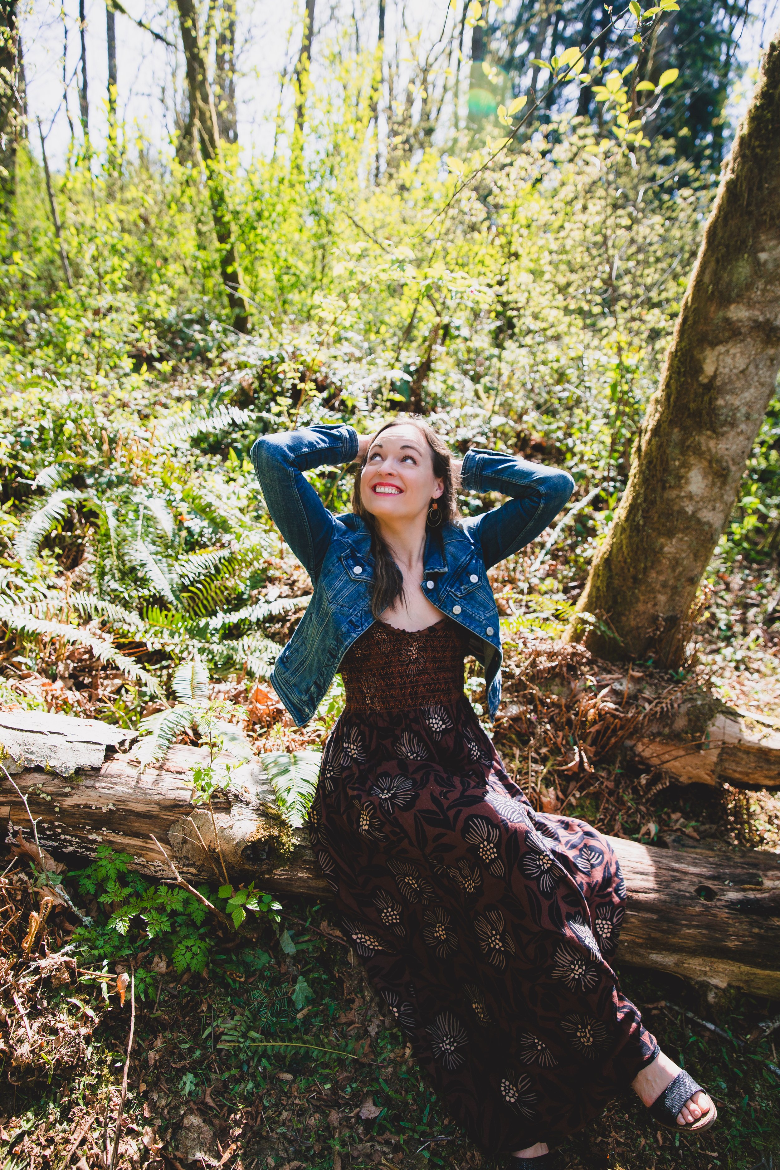 A woman sitting on a fallen log in a forest, looking up with a smile, surrounded by green foliage and sunlight filtering through the trees.