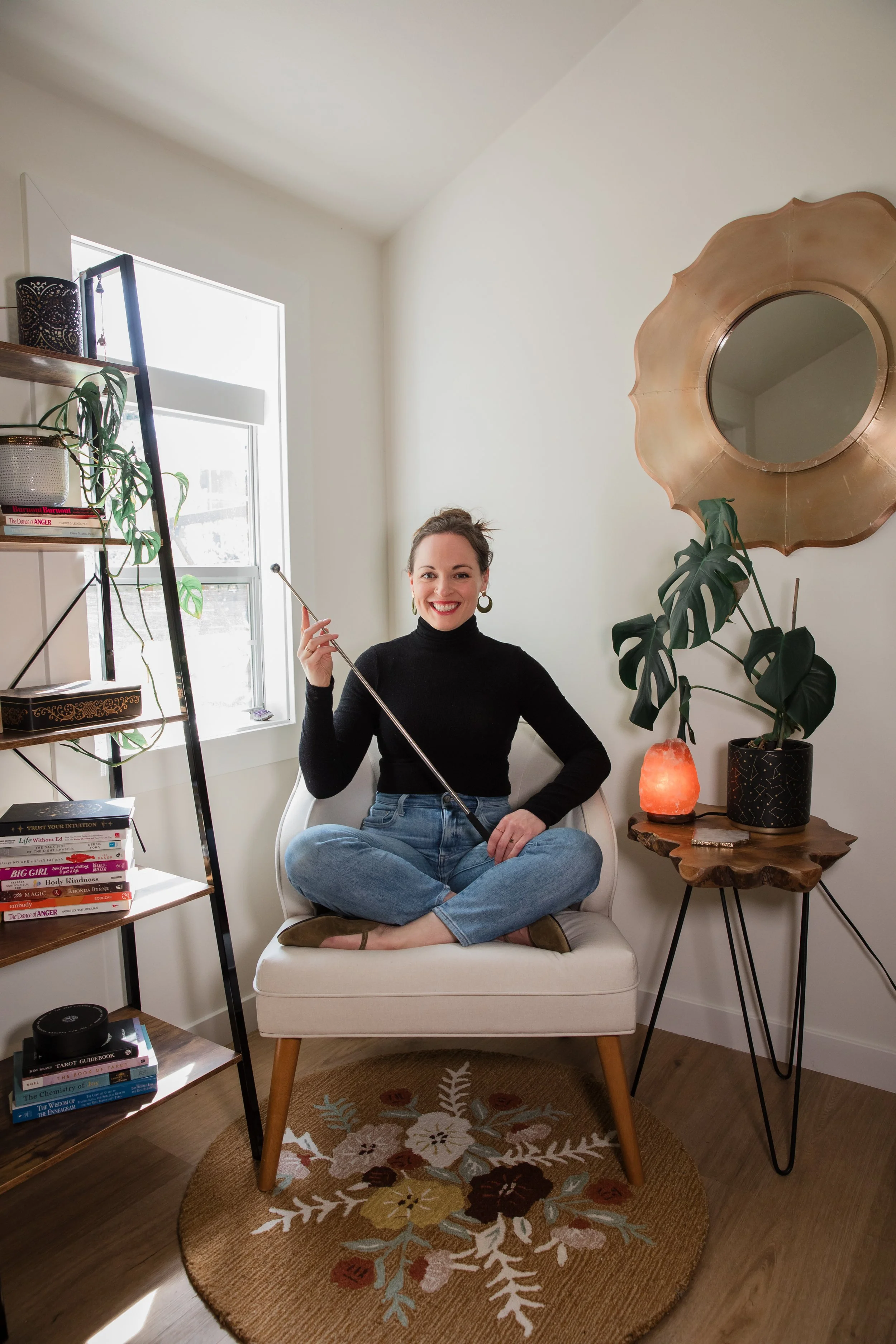 A woman sitting cross-legged on a cream-colored armchair, holding a pointer, smiling in a cozy room with books, plants, a salt lamp, a large decorative mirror, and a round patterned rug.
