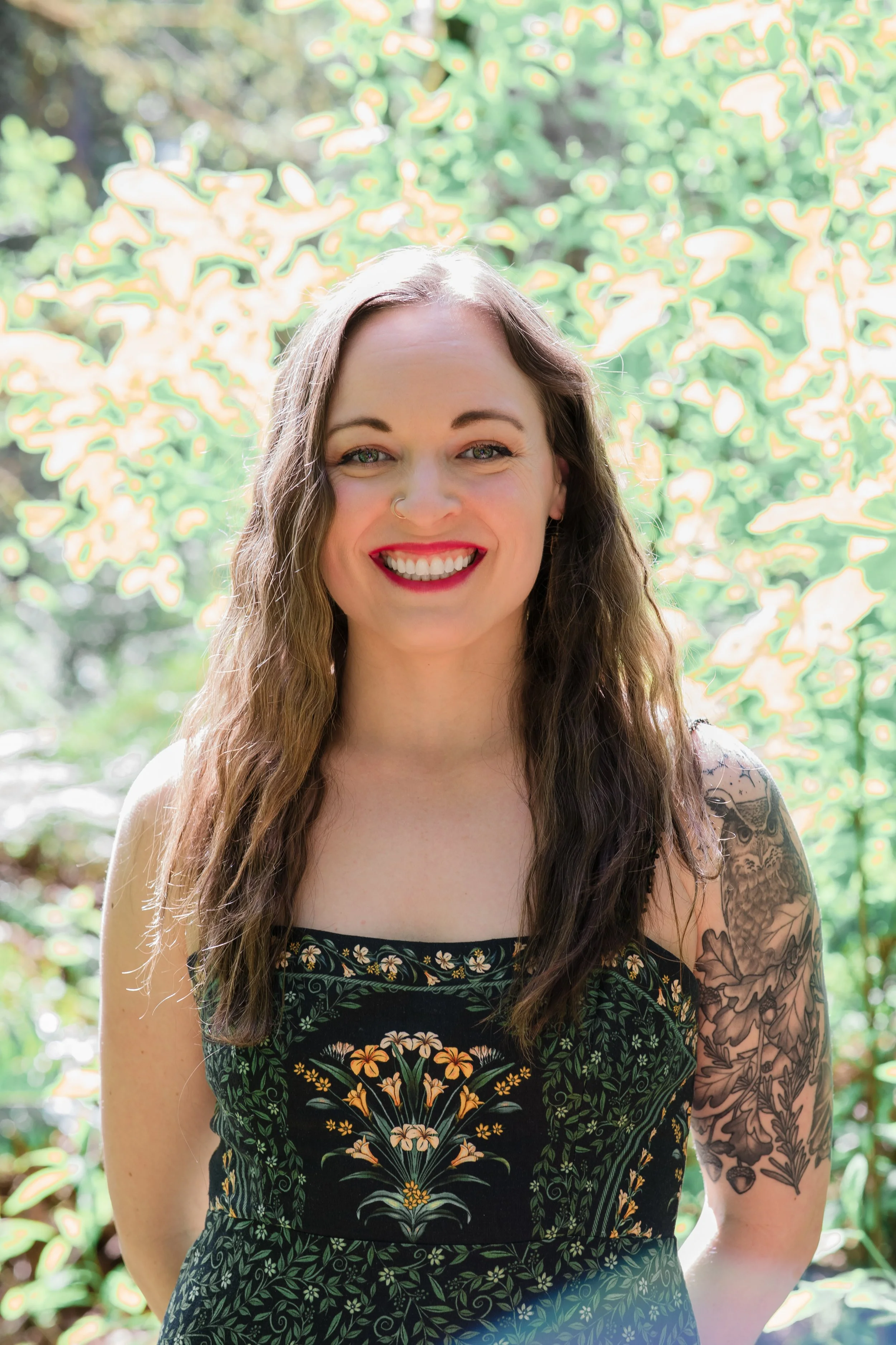 Young woman with long brown hair and tattoos on her arm, smiling outdoors in a sunny setting with green trees in the background.