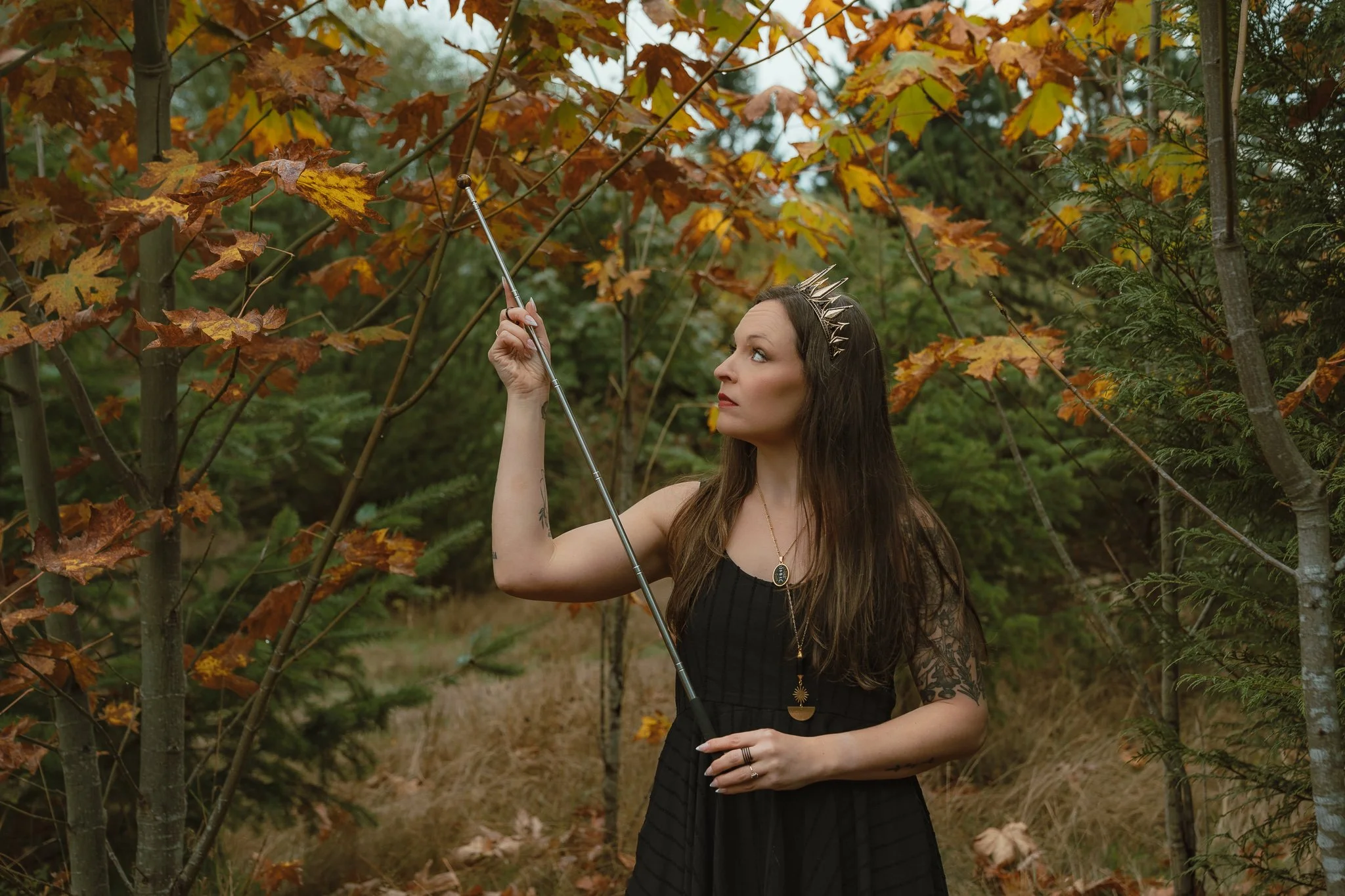 A woman wearing a black dress and a spiked crown is outdoors, holding a small portable mirror near autumn leaves on a tree, with trees and grass in the background.
