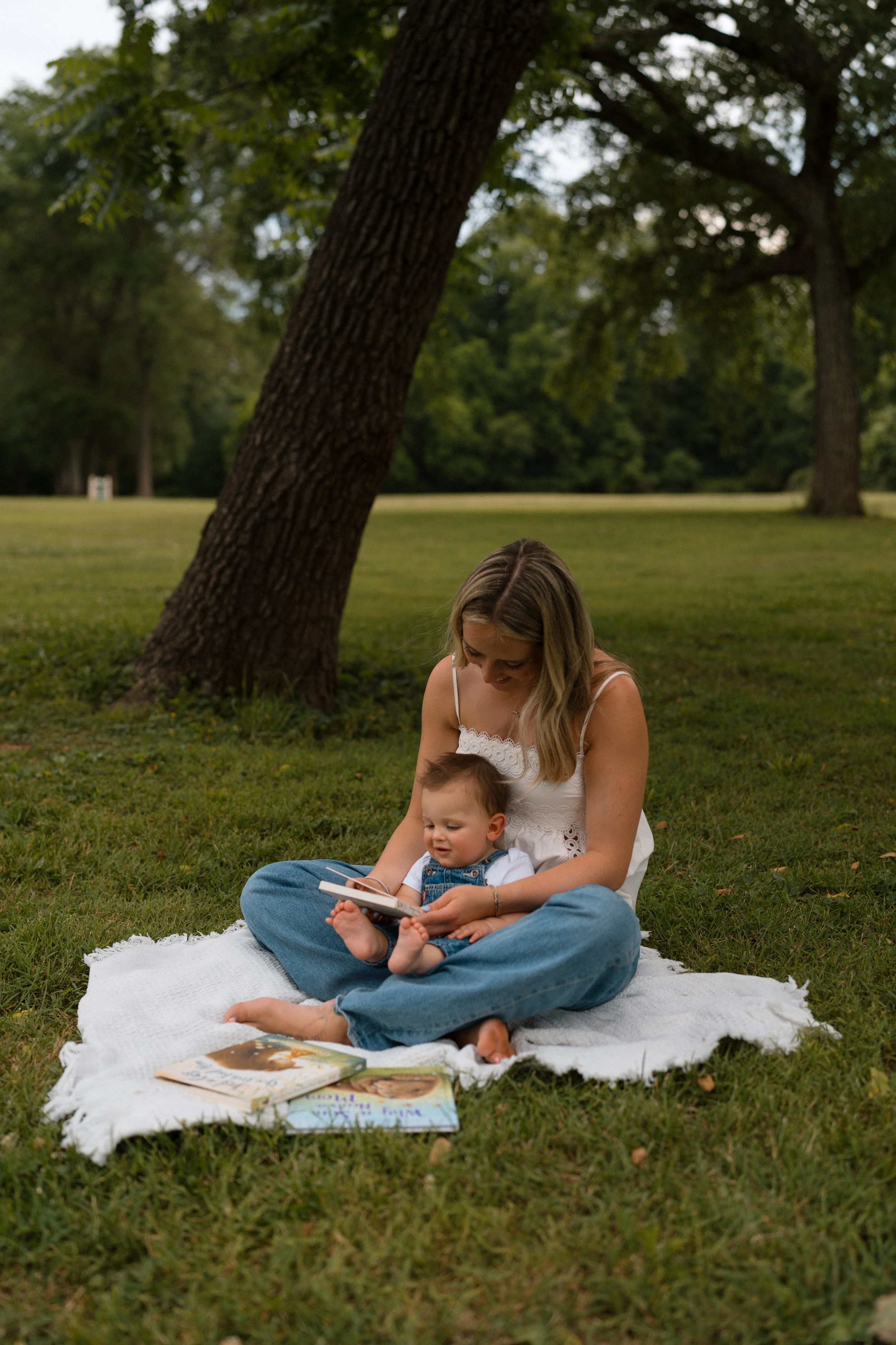 A woman and a young child sitting on a white blanket on the grass in a park. The woman is holding the child and they are looking at a book. Multiple books are spread out on the blanket, and there are large trees in the background.